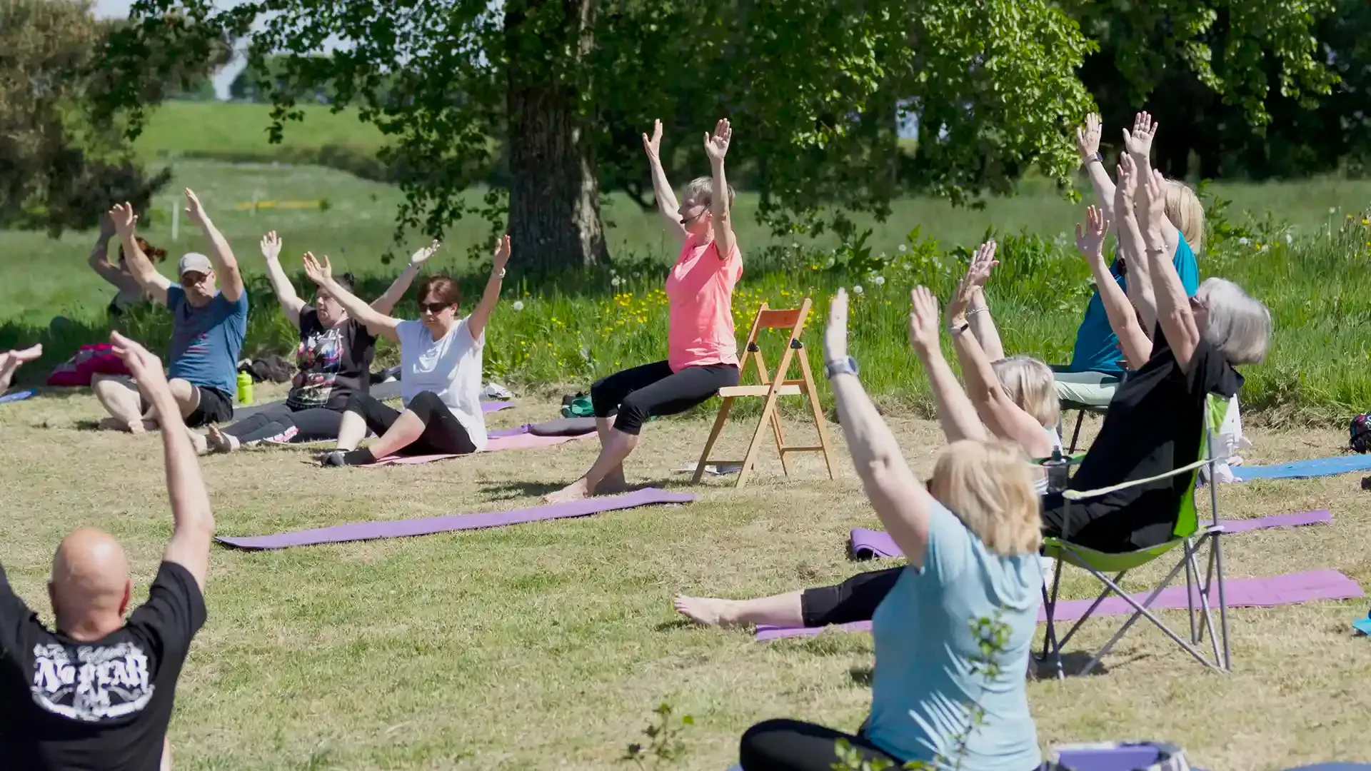 People practising yoga outdoors, some seated on the ground and some seated-on chairs.