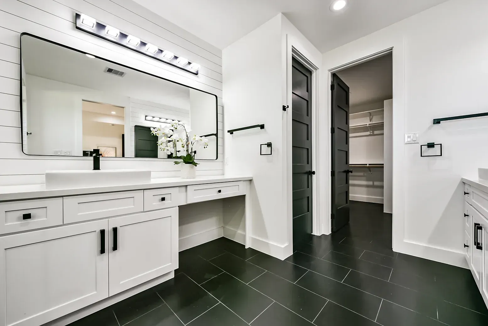 A bathroom with black tile floors , white cabinets and a large mirror.