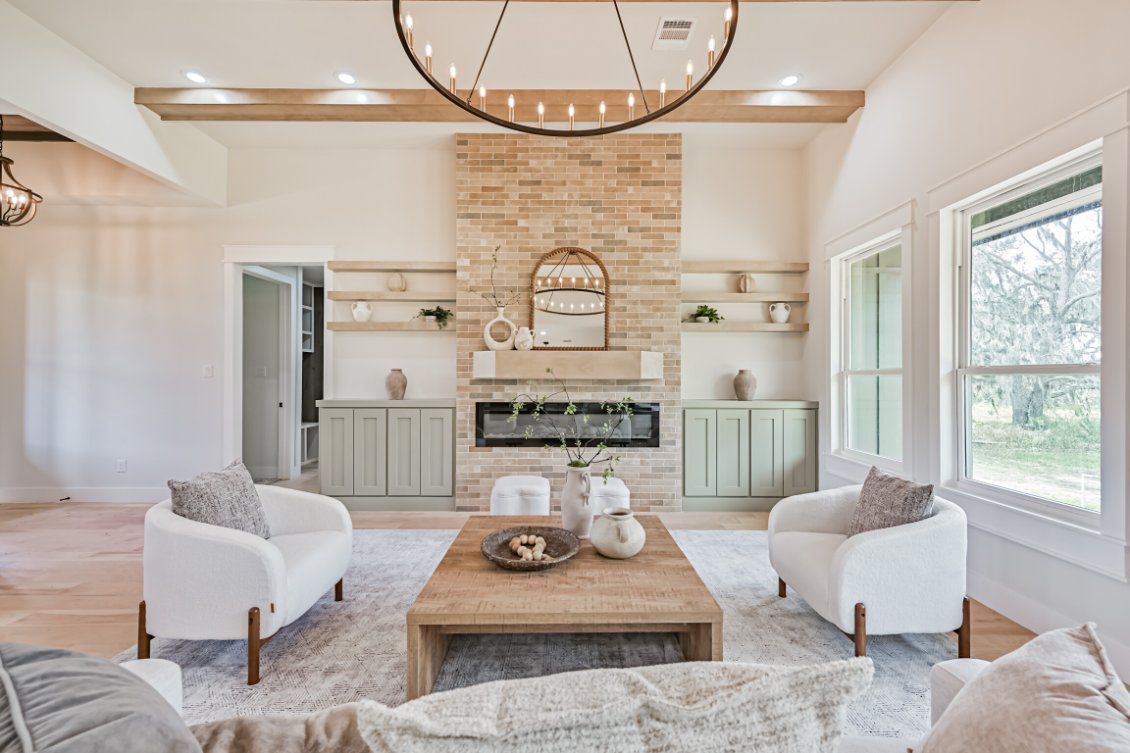 A bright living room with a stone fireplace, light wood coffee table, two white armchairs, and a large metal chandelier.