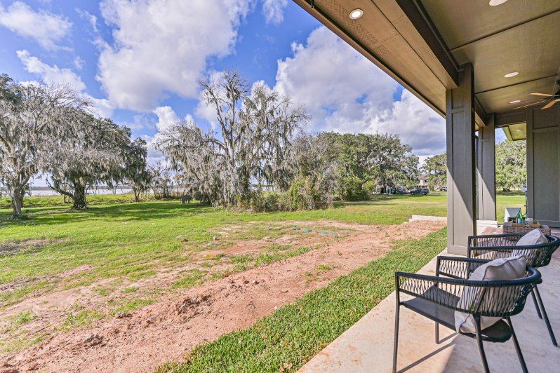 A covered patio with two black chairs looks out over a grassy yard with trees under a blue sky with fluffy white clouds.