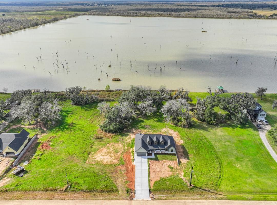 An aerial view of a lakefront property featuring a newly constructed house, green lawn, and scattered mature trees.