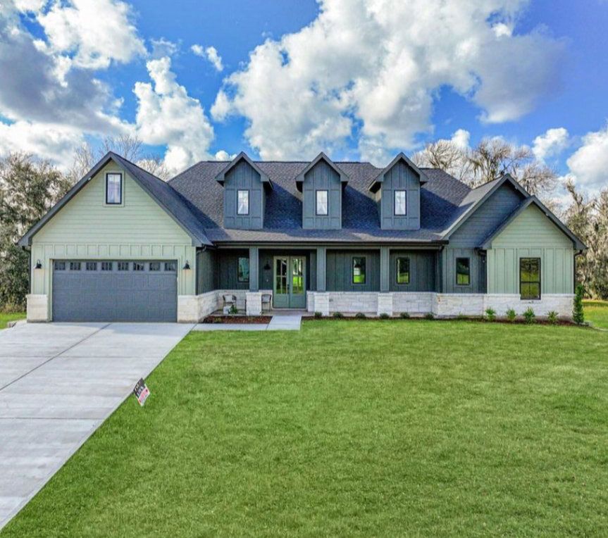 A single-story suburban house with sage green siding, dark gray roof, and white stone base, fronted by a large grassy lawn.