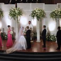 Wedding ceremony on a stage with bride, groom, officiant, and wedding party before big white floral backdrops