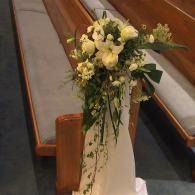 White and green floral arrangement attched at the end of a wooden church pew bench
