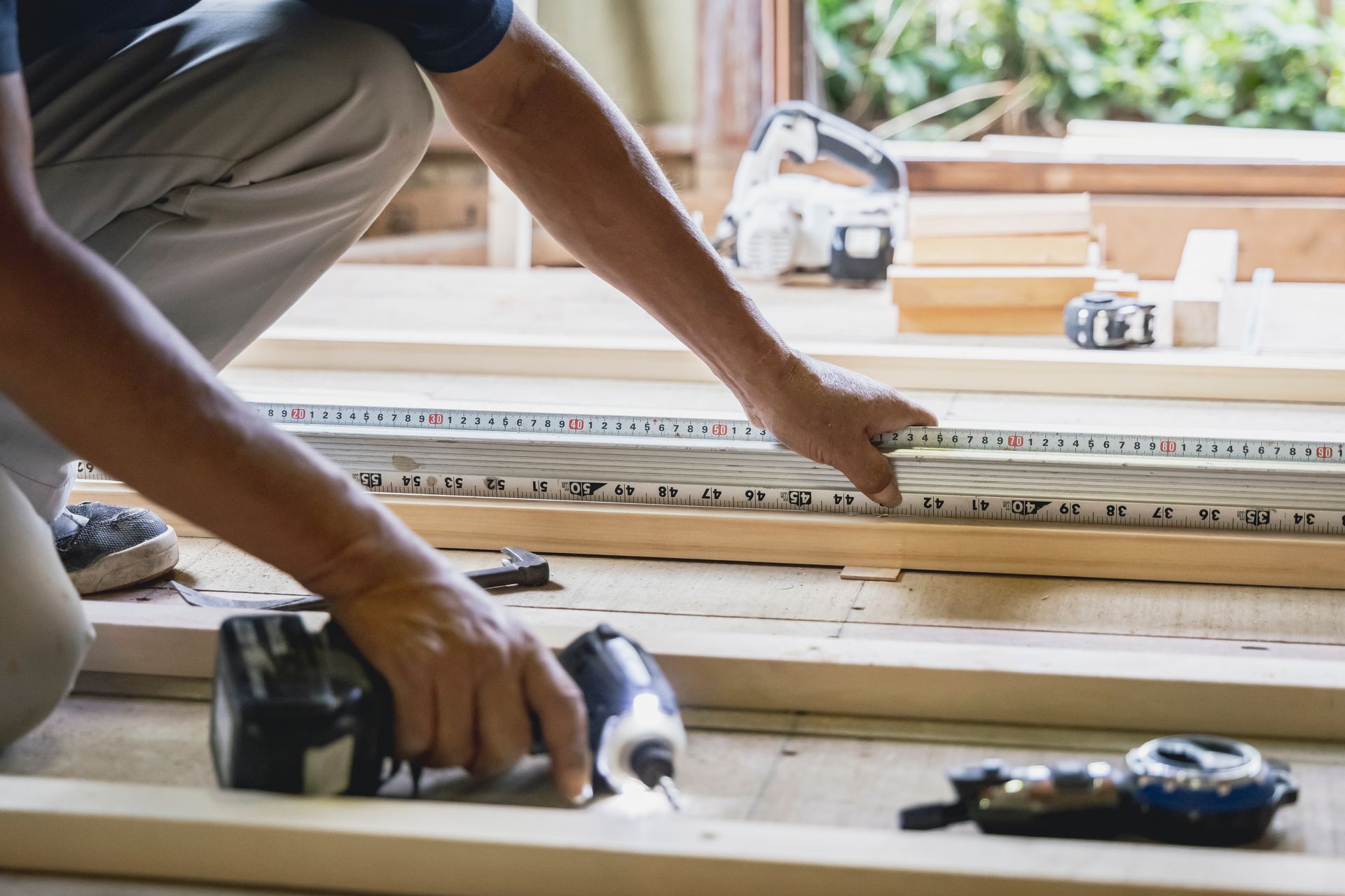 A person kneels on wooden floor joists, using a power drill to fasten a measuring tool against a wooden board.