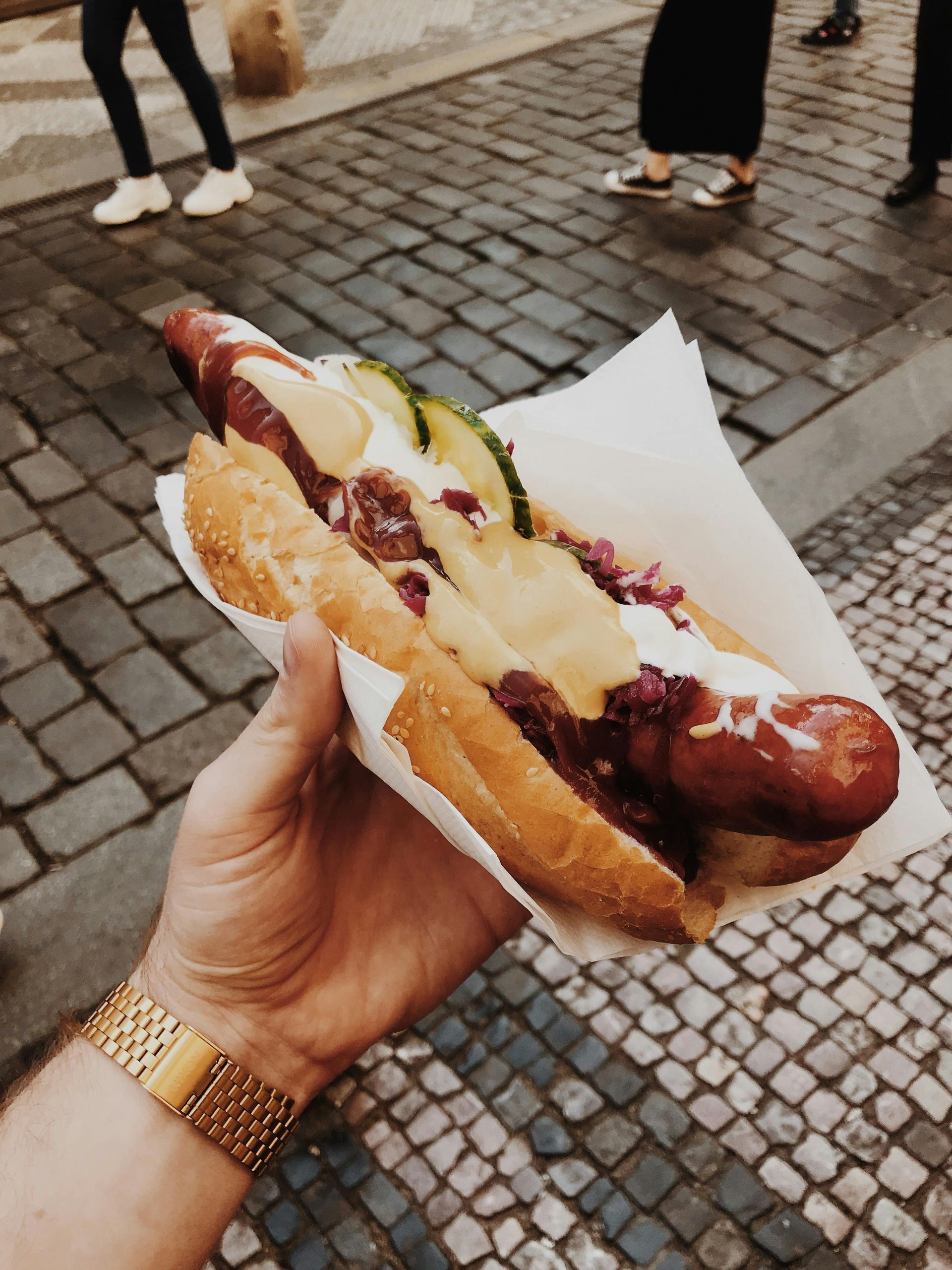 Eine Person hält einen Hotdog auf einem gepflasterten Gehweg.