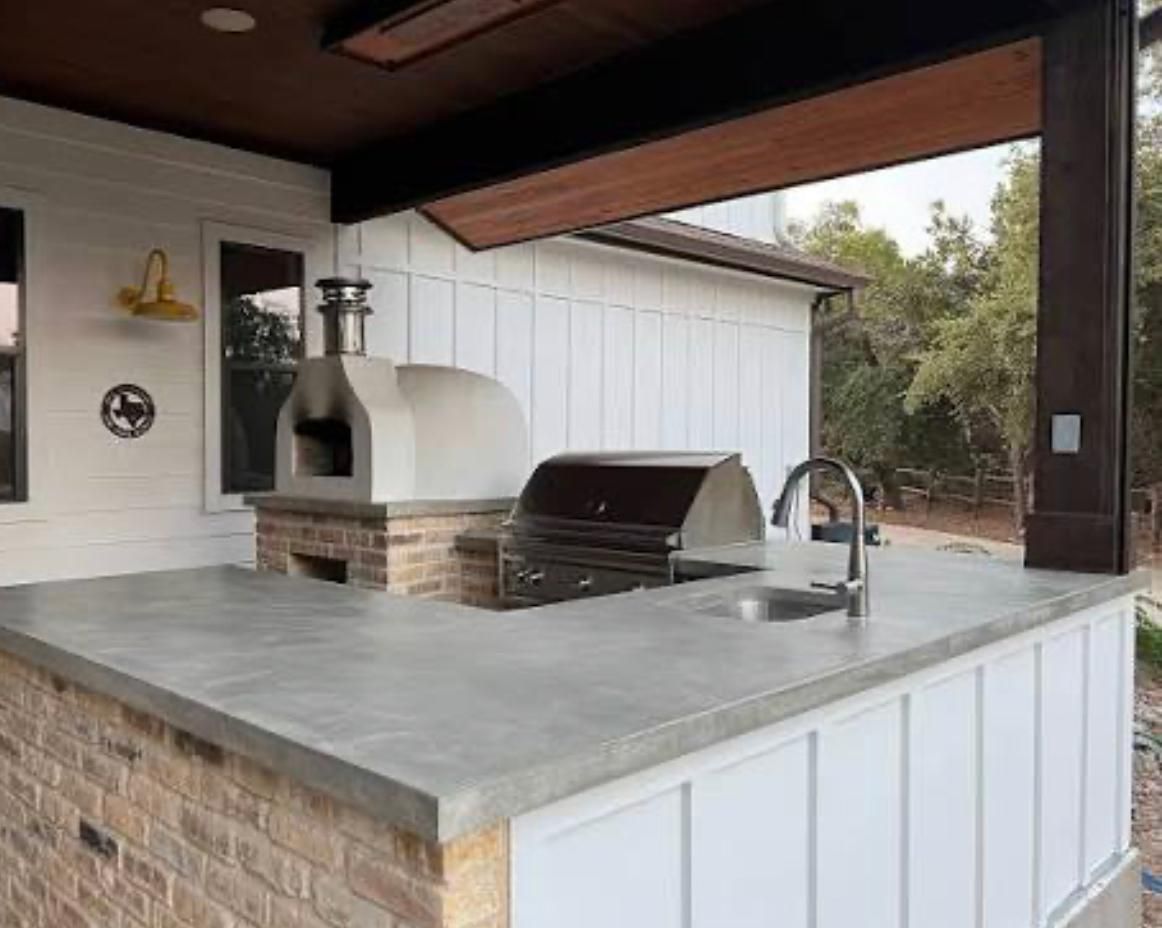 Outdoor kitchen with a concrete countertop, brick base, pizza oven, grill, and sink on a covered patio.