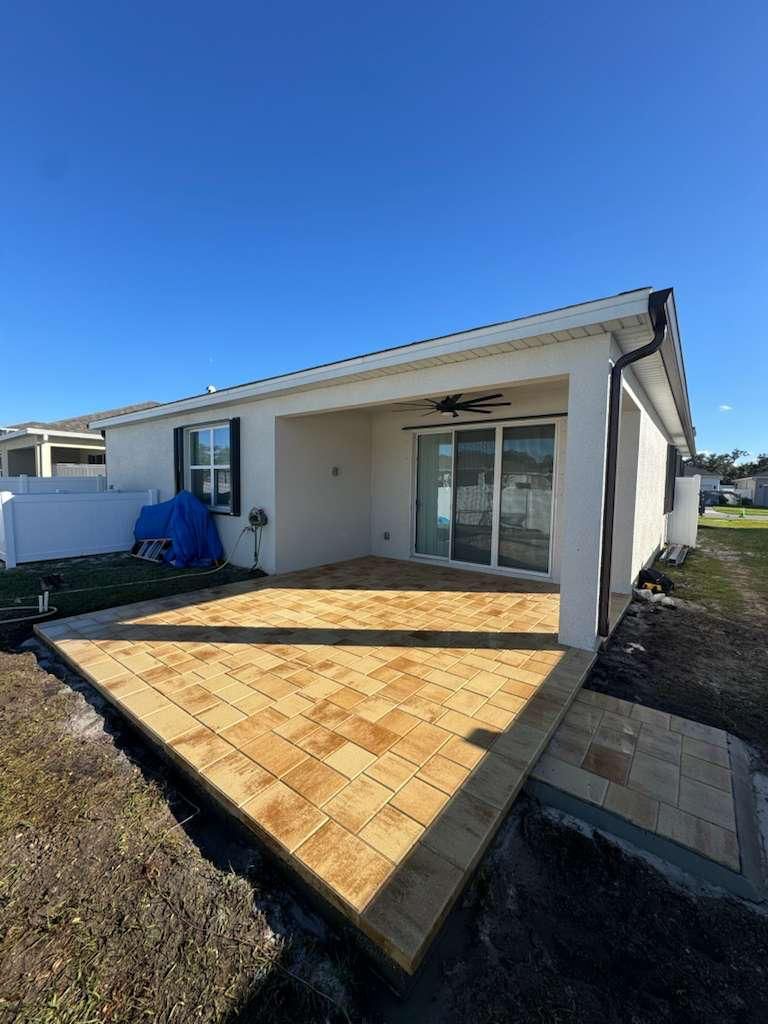 A sunny outdoor patio features tan pavers extending from a covered white house area with sliding glass doors.