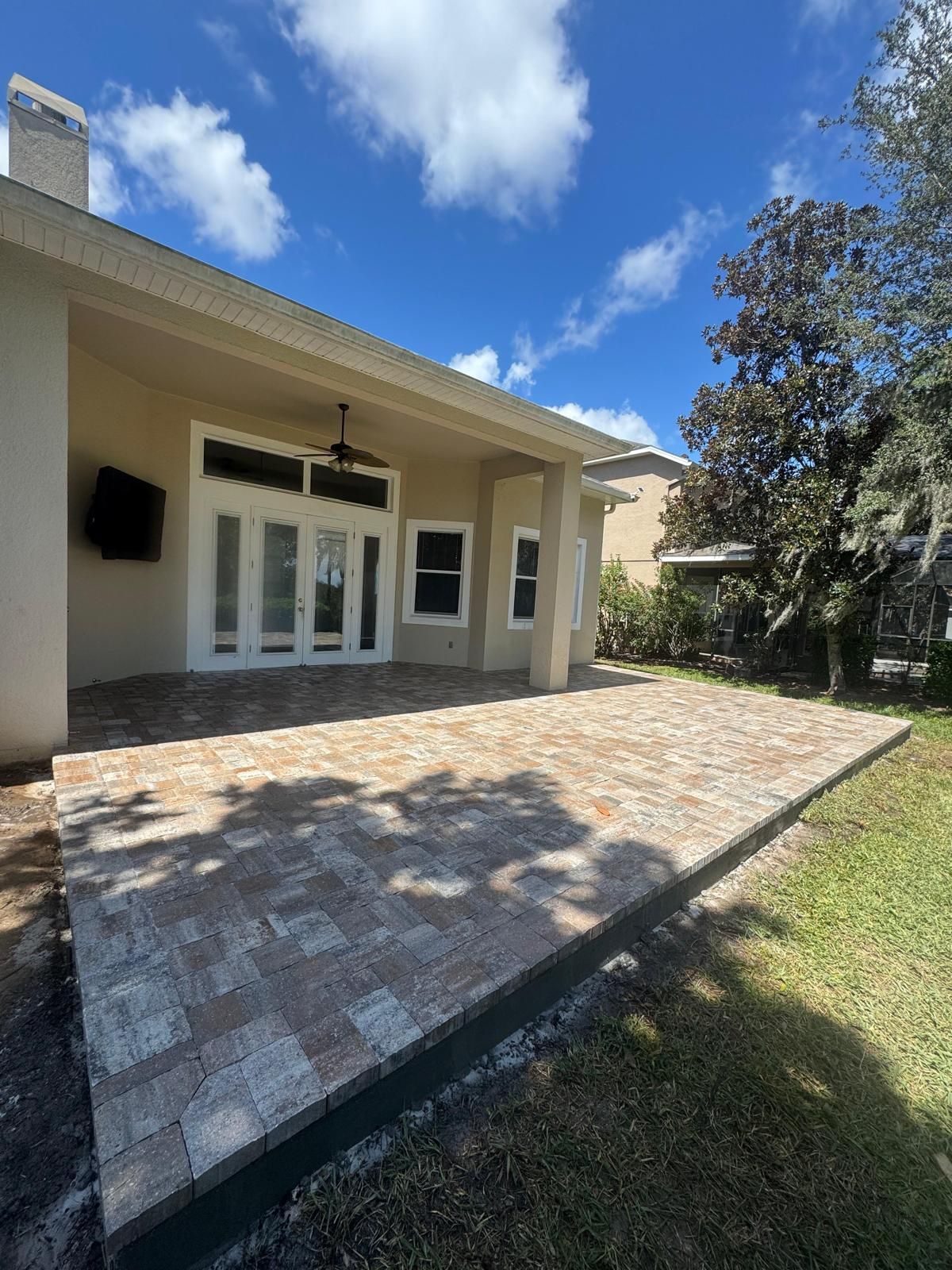 A beige house with a covered patio featuring a newly laid, multi-toned stone paver floor, next to a large green tree.