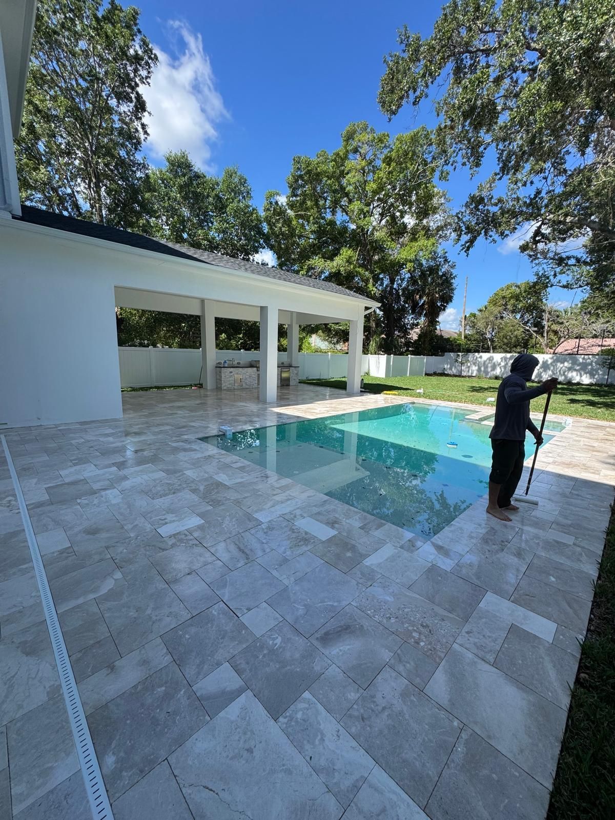 A person cleans a poolside patio made of light stone tiles next to a covered outdoor living area on a sunny day.