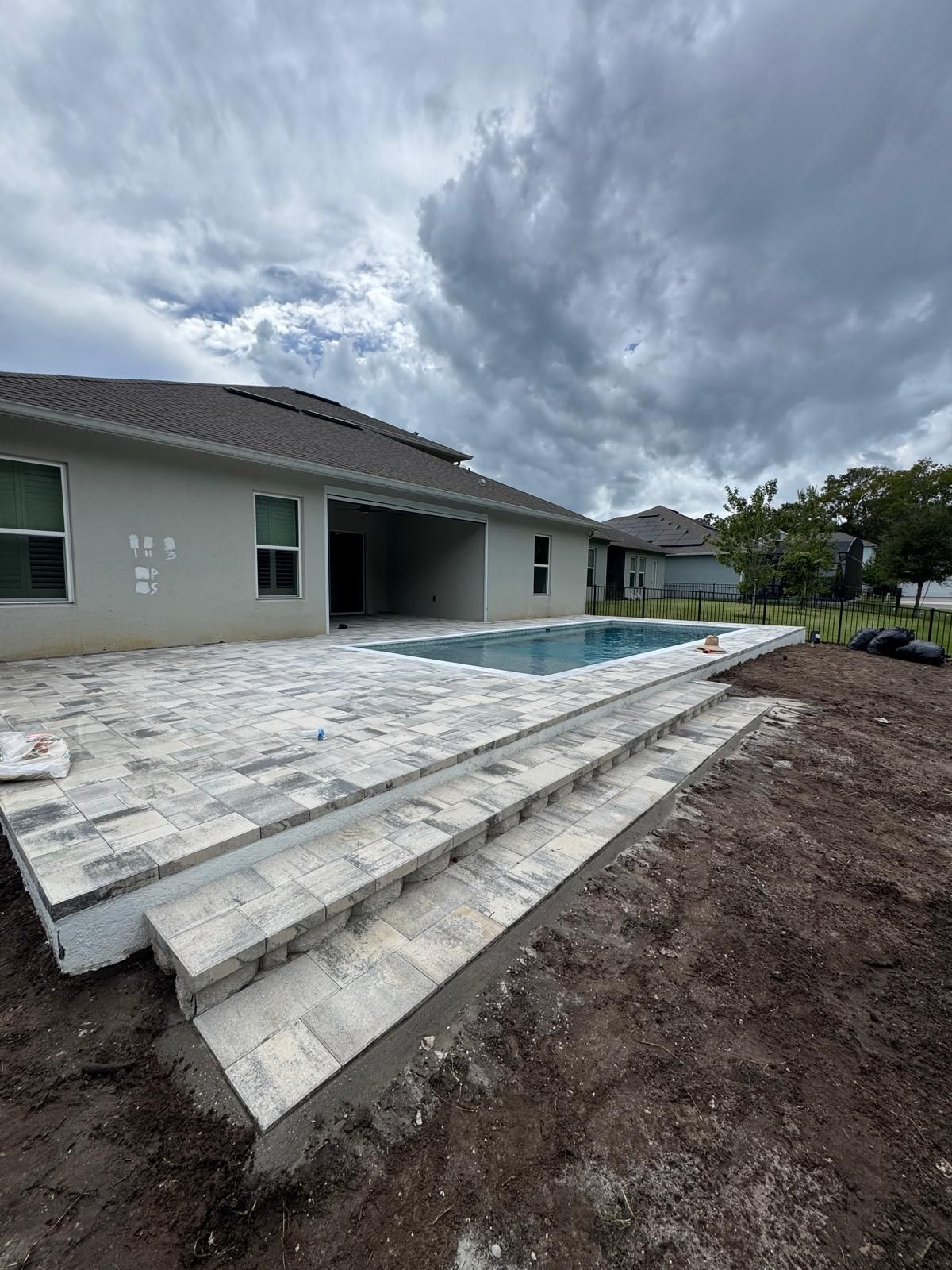 A gray house with a stone paver patio and rectangular swimming pool, set against a cloudy, overcast sky.
