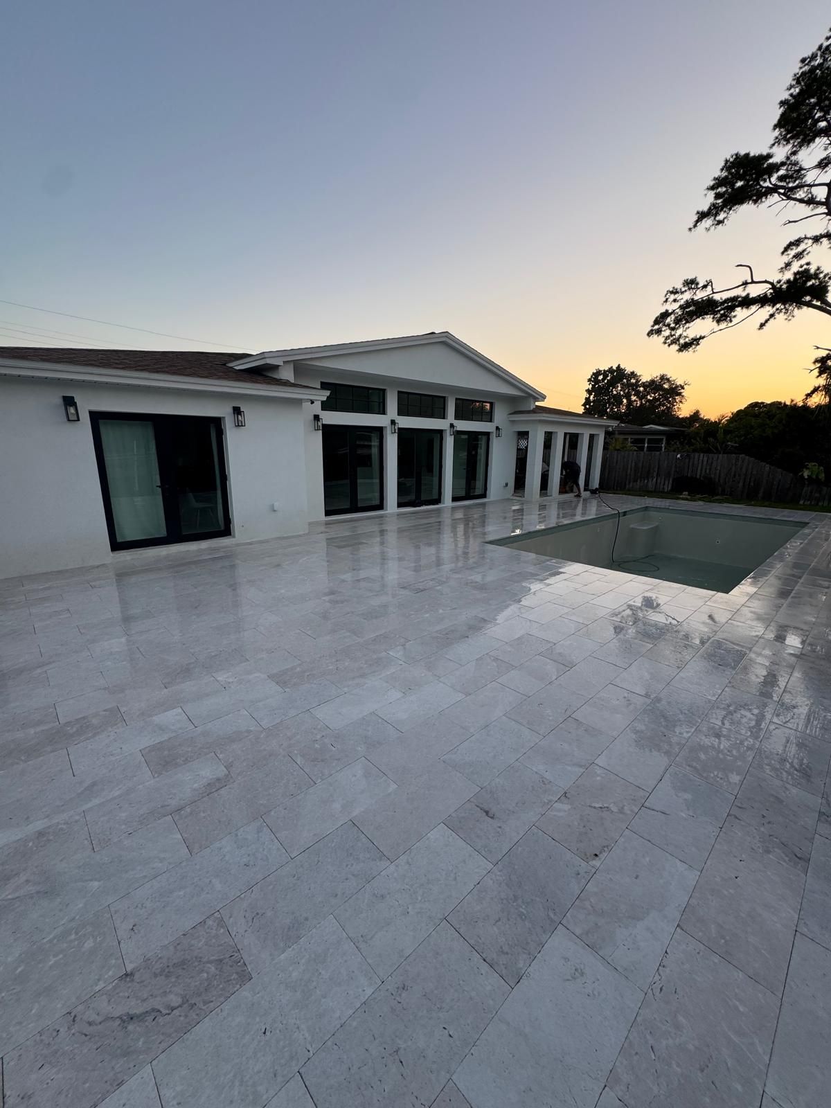 A white house with a large stone patio and an empty swimming pool at sunset.