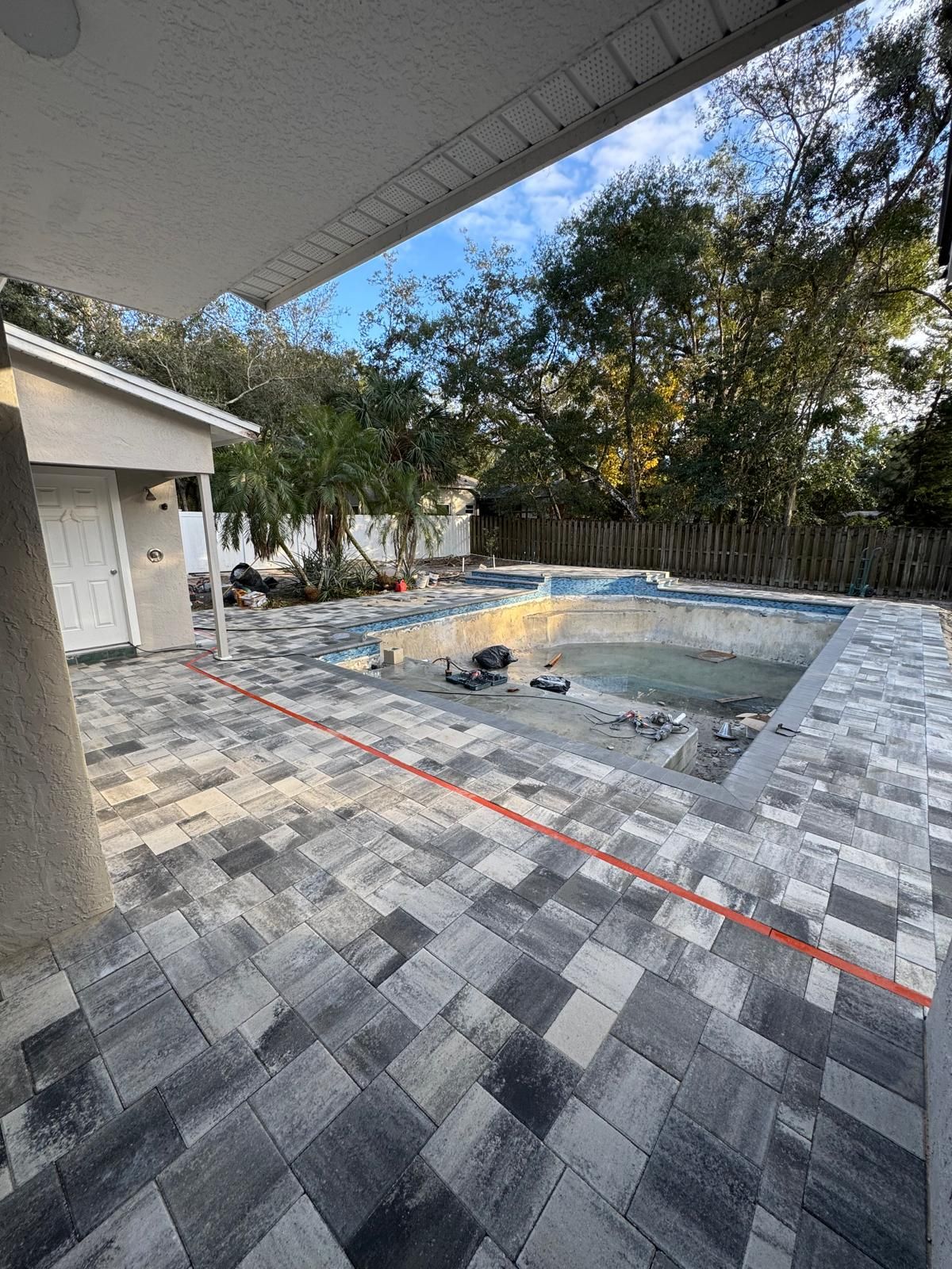 A patio paved with grey stone tiles surrounds an uncompleted swimming pool with a red construction line on the ground.