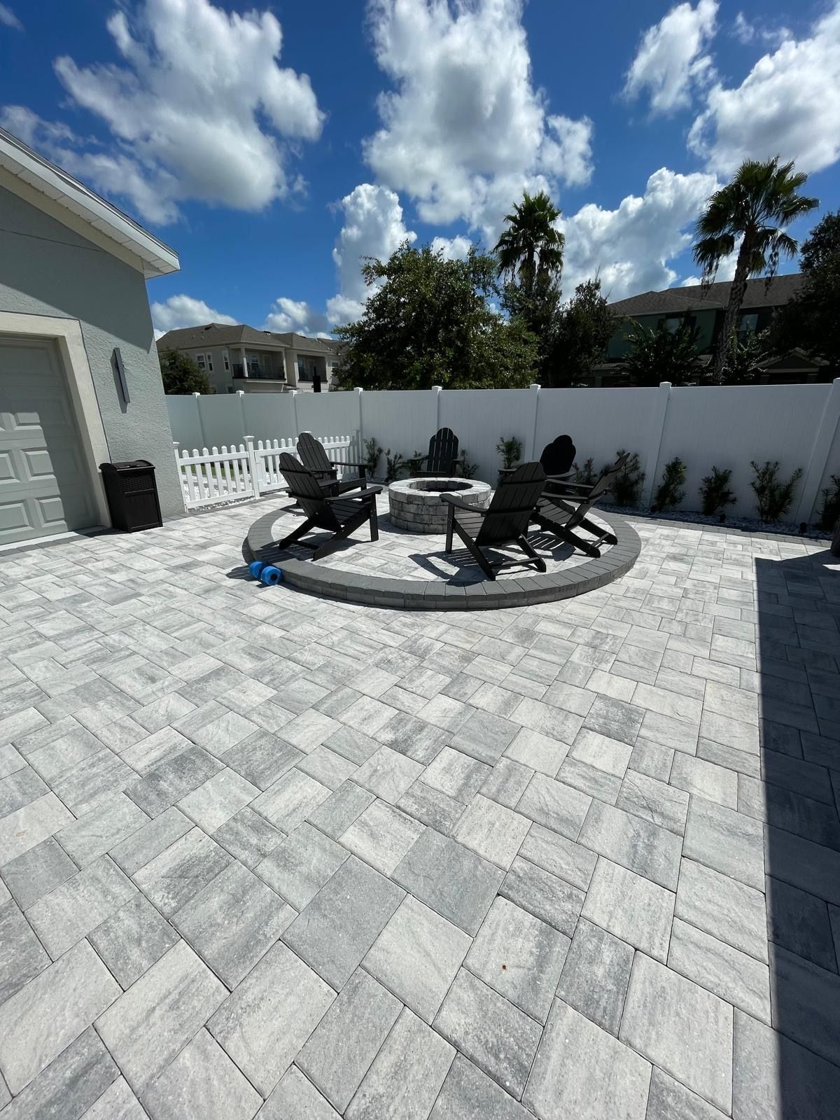 Patio with gray stone pavers, a circular fire pit, and black adirondack chairs, enclosed by a white fence under blue skies.