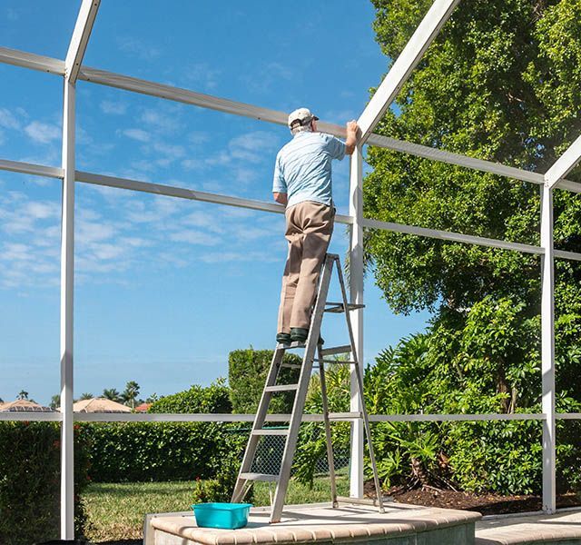 Man on ladder, repairing white screen structure against a blue sky with green foliage.