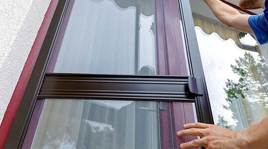 Brown screen door being installed on a white building, with a person's hands visible.