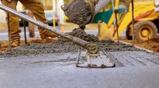 Concrete being poured from a truck, smoothed by a worker with a large trowel in a construction setting.
