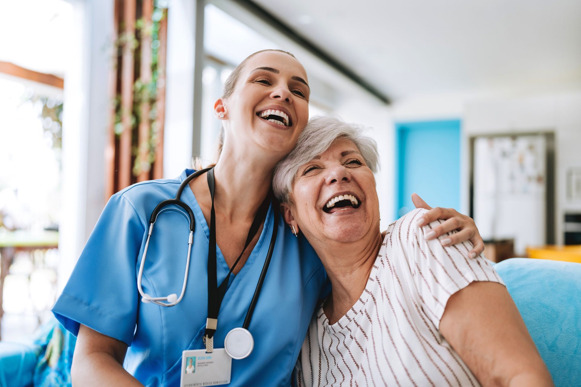 A nurse and an elderly woman are laughing together while sitting on a couch