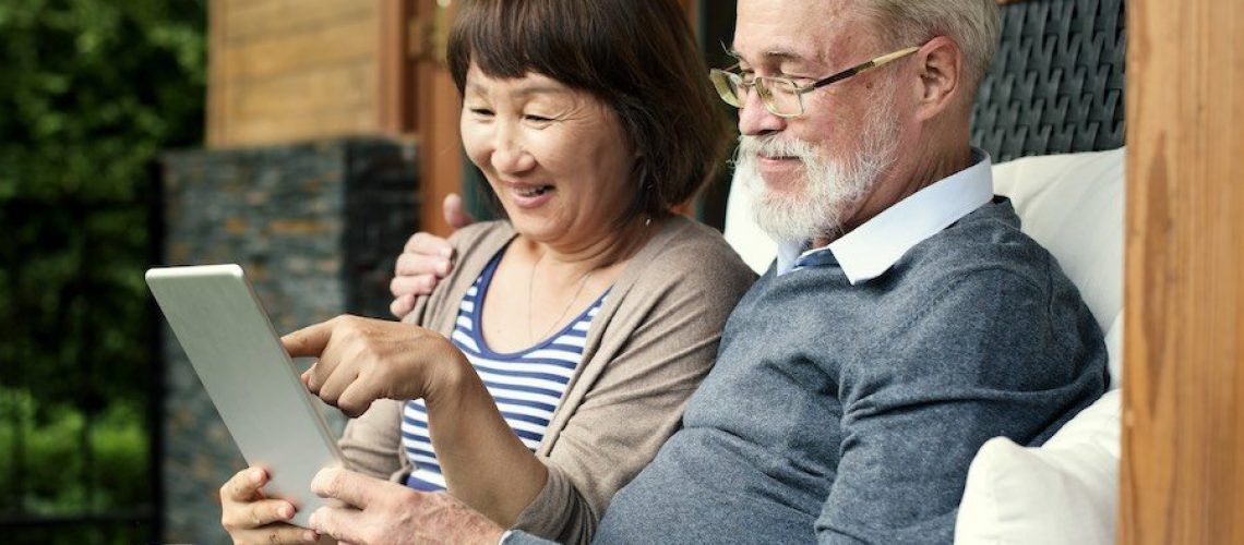 An elderly couple is sitting on a couch looking at a tablet.