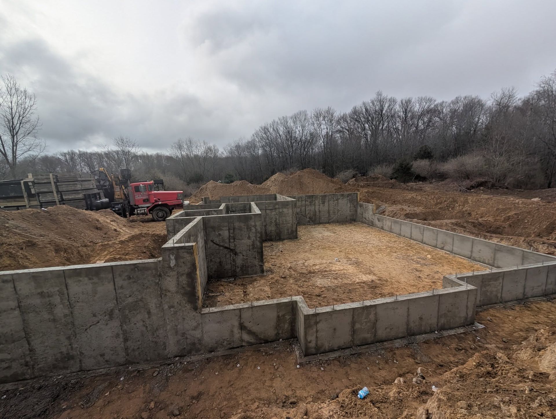 A concrete foundation for a new building under construction on a dirt lot with trees in the background under a cloudy sky.