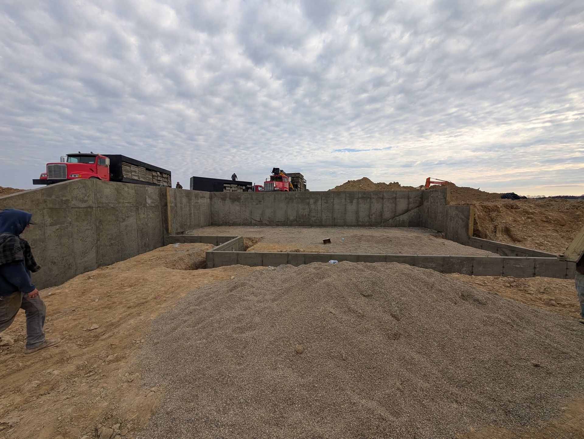 A partially built concrete foundation for a building site with piles of gravel and construction trucks in the background.