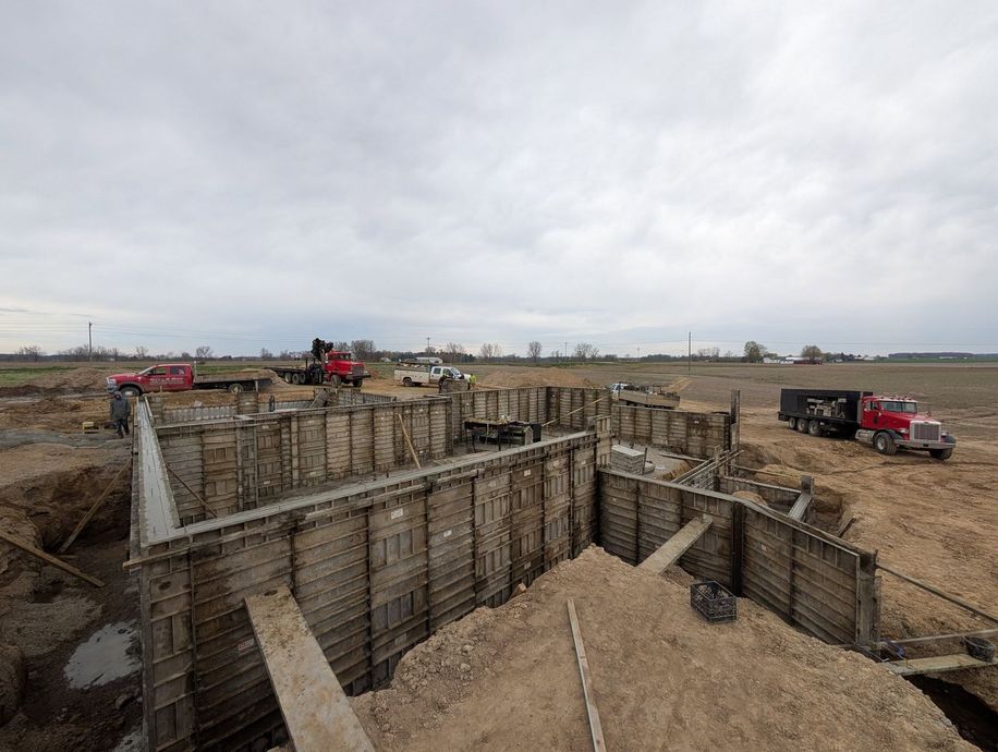 Concrete foundation forms for a new building under construction in a flat, rural field under a cloudy sky.