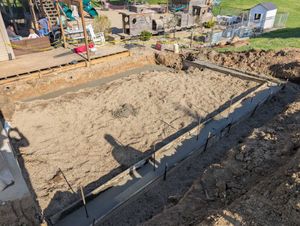 Freshly poured concrete footings for a building project set in a dirt-filled, excavated rectangle in a backyard.