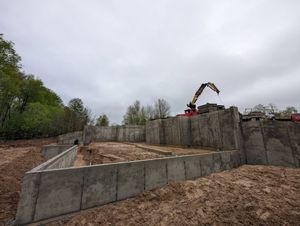 A construction site features concrete foundation walls and an excavator working on a cloudy day.