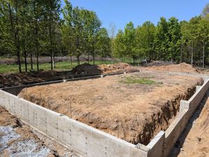 Concrete foundation walls enclose a rectangular plot of dirt in a rural, wooded setting under a clear blue sky.