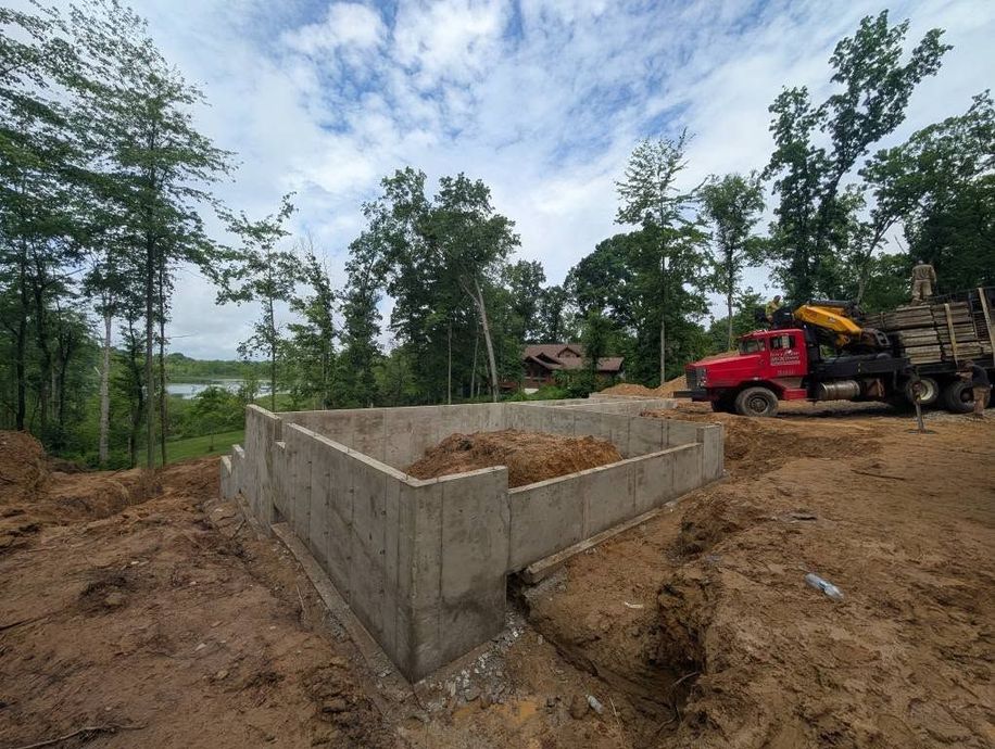 A concrete foundation for a new house under construction in a wooded area with a red truck parked nearby.