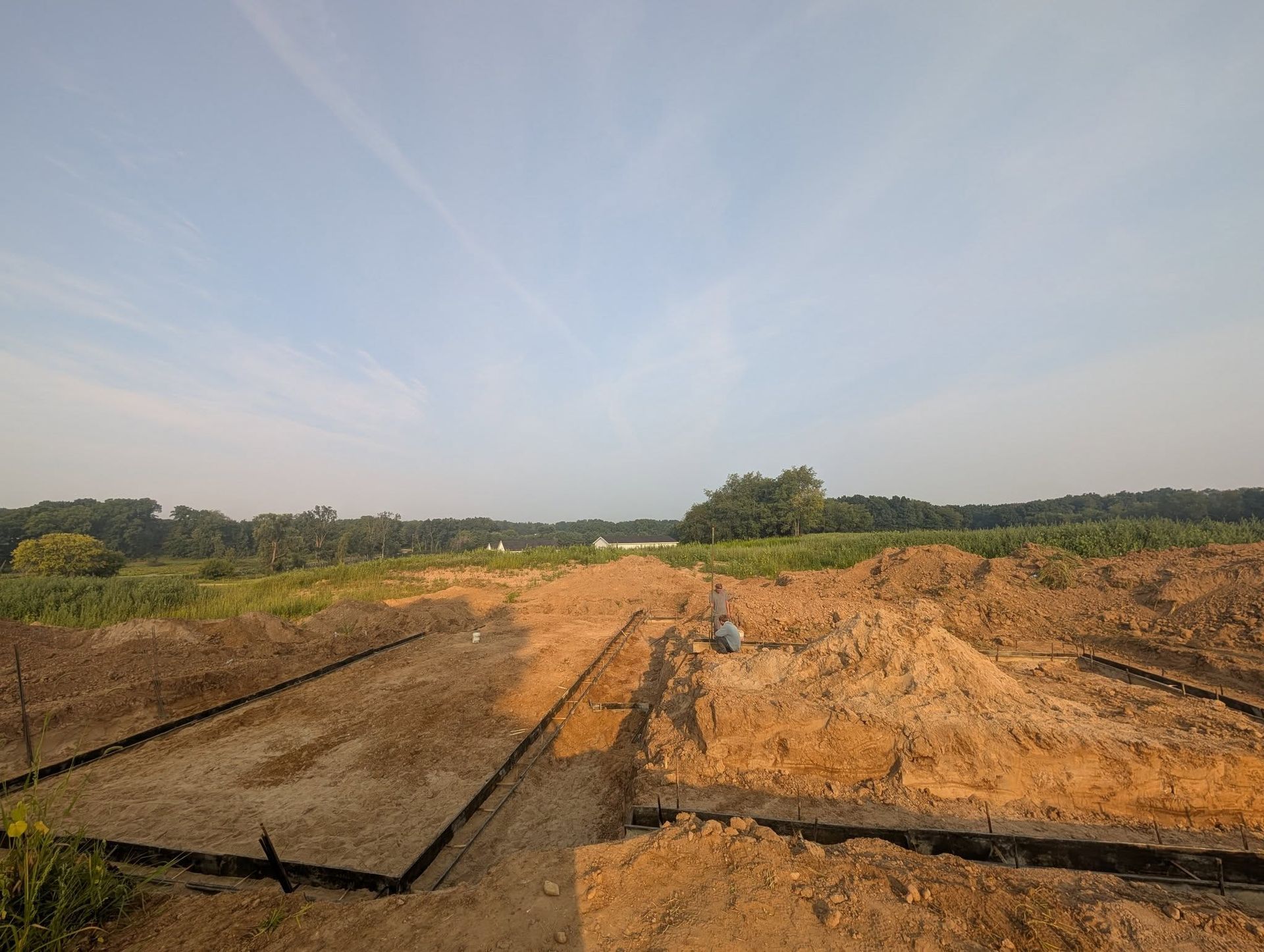 A construction site showing trenches dug into the ground with dark liners, surrounded by piles of dirt under a clear sky.
