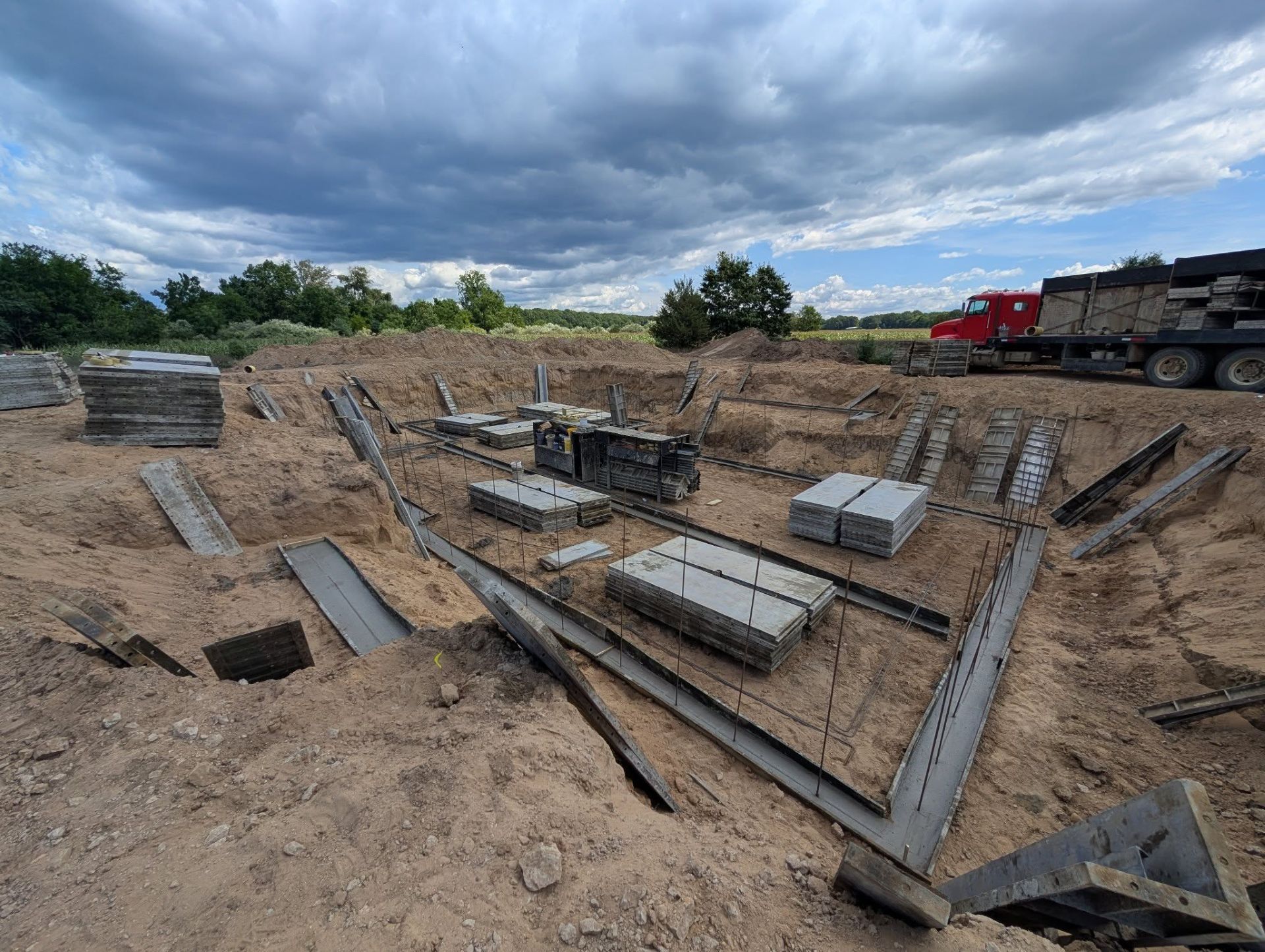 A construction site with concrete foundation forms, steel beams, and stacks of blocks in a dirt excavation under a cloudy sky.