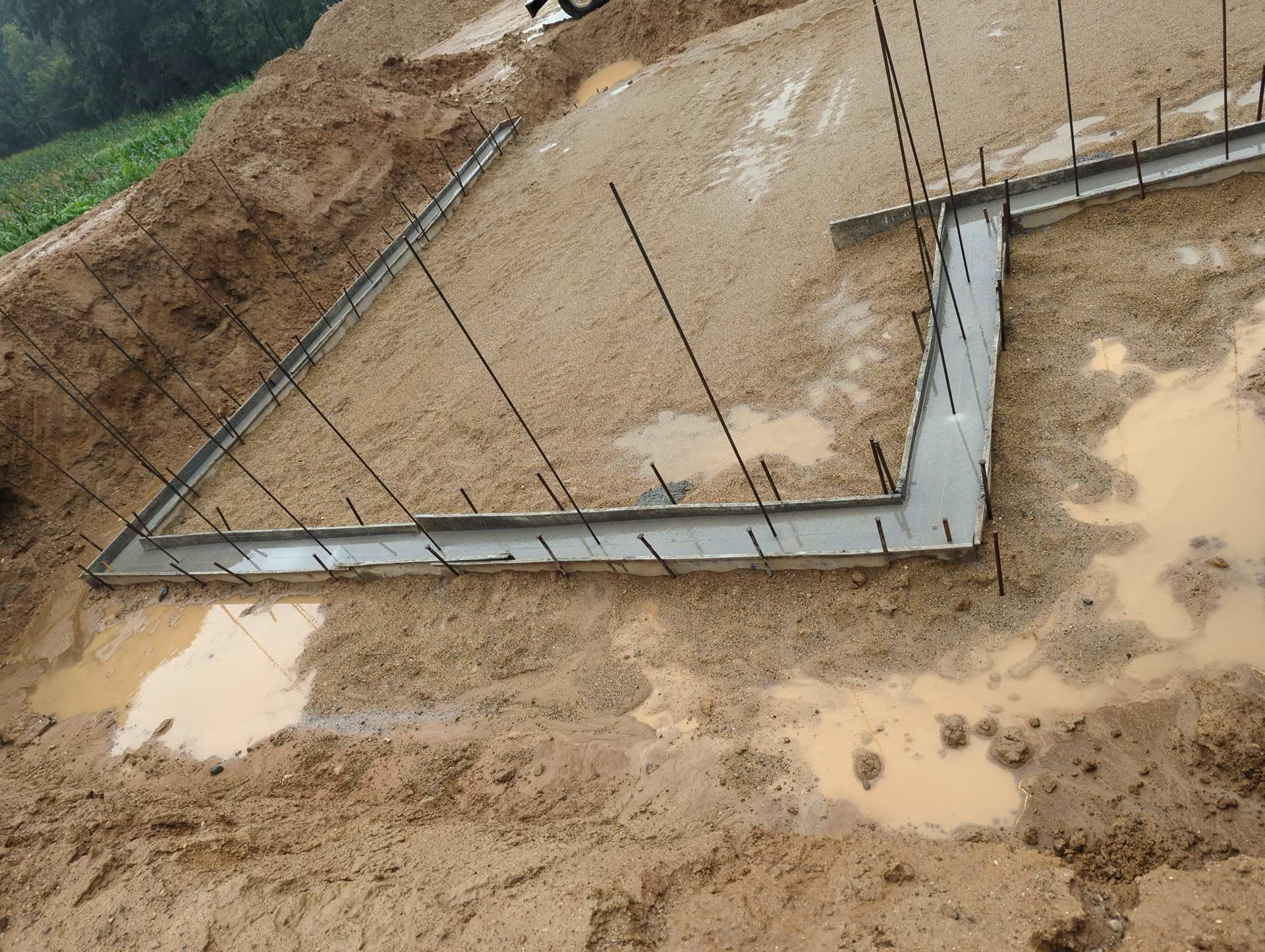 A concrete foundation with upright rebar rods set in a muddy construction site.