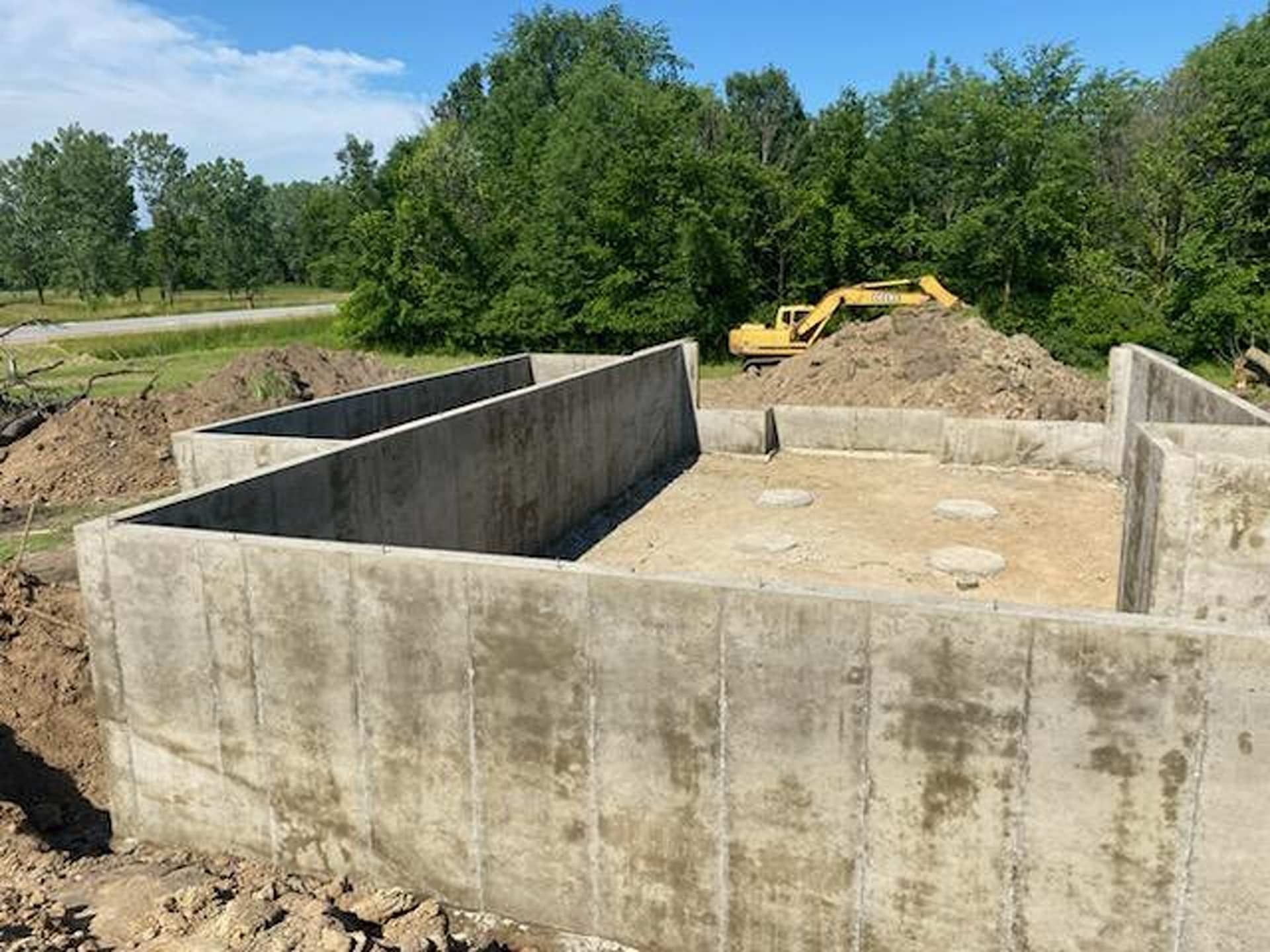 A poured concrete foundation for a new house construction, surrounded by mounds of dirt and trees under a clear blue sky.