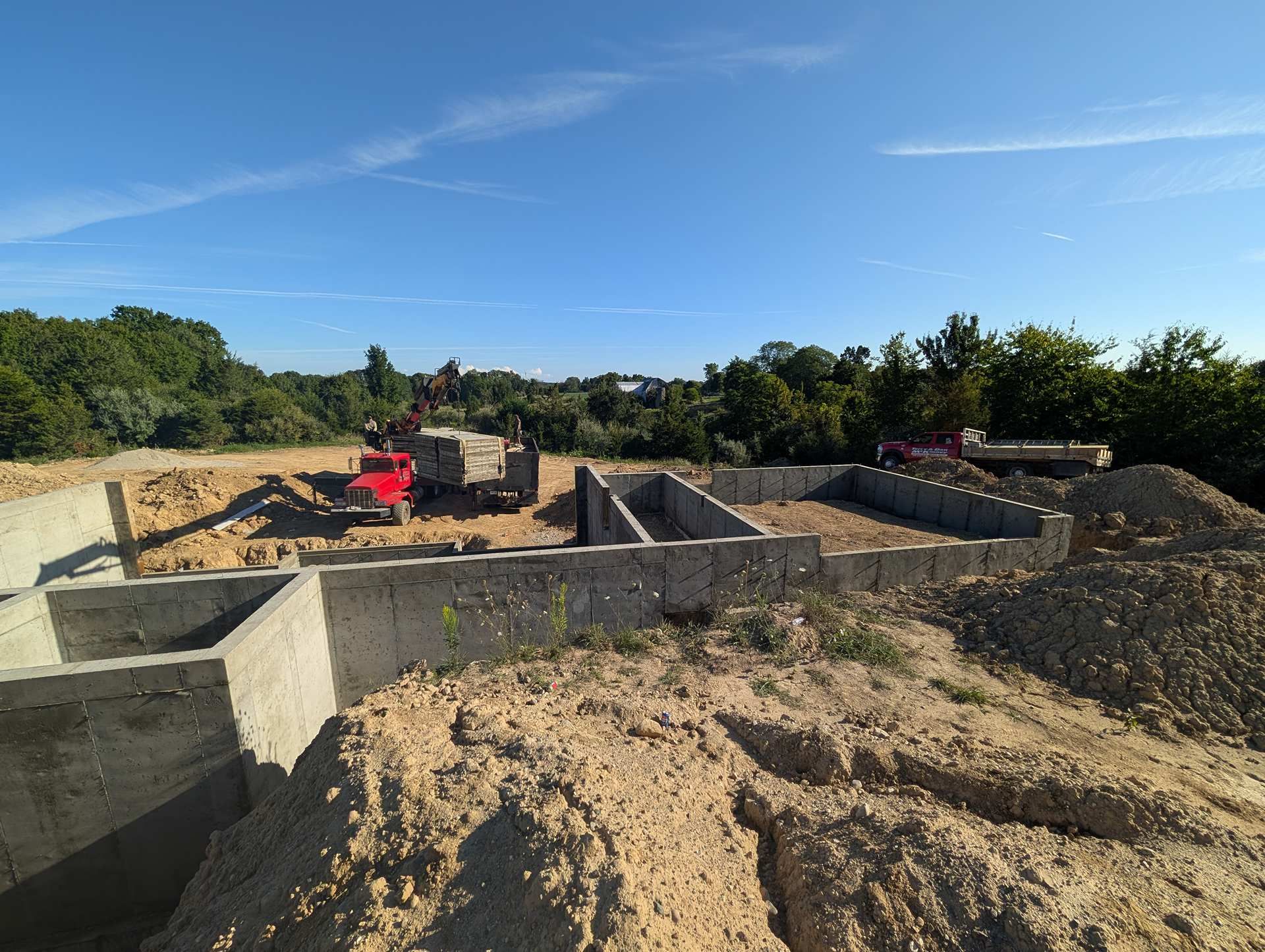Concrete foundation walls of a building under construction on a sunny day, with piles of dirt and a red truck nearby.