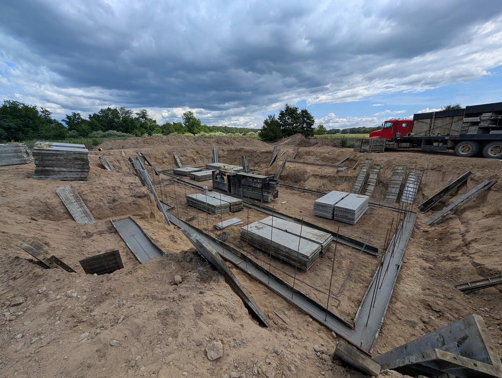 A construction site showing concrete foundations and footings in a dirt lot under a cloudy sky.