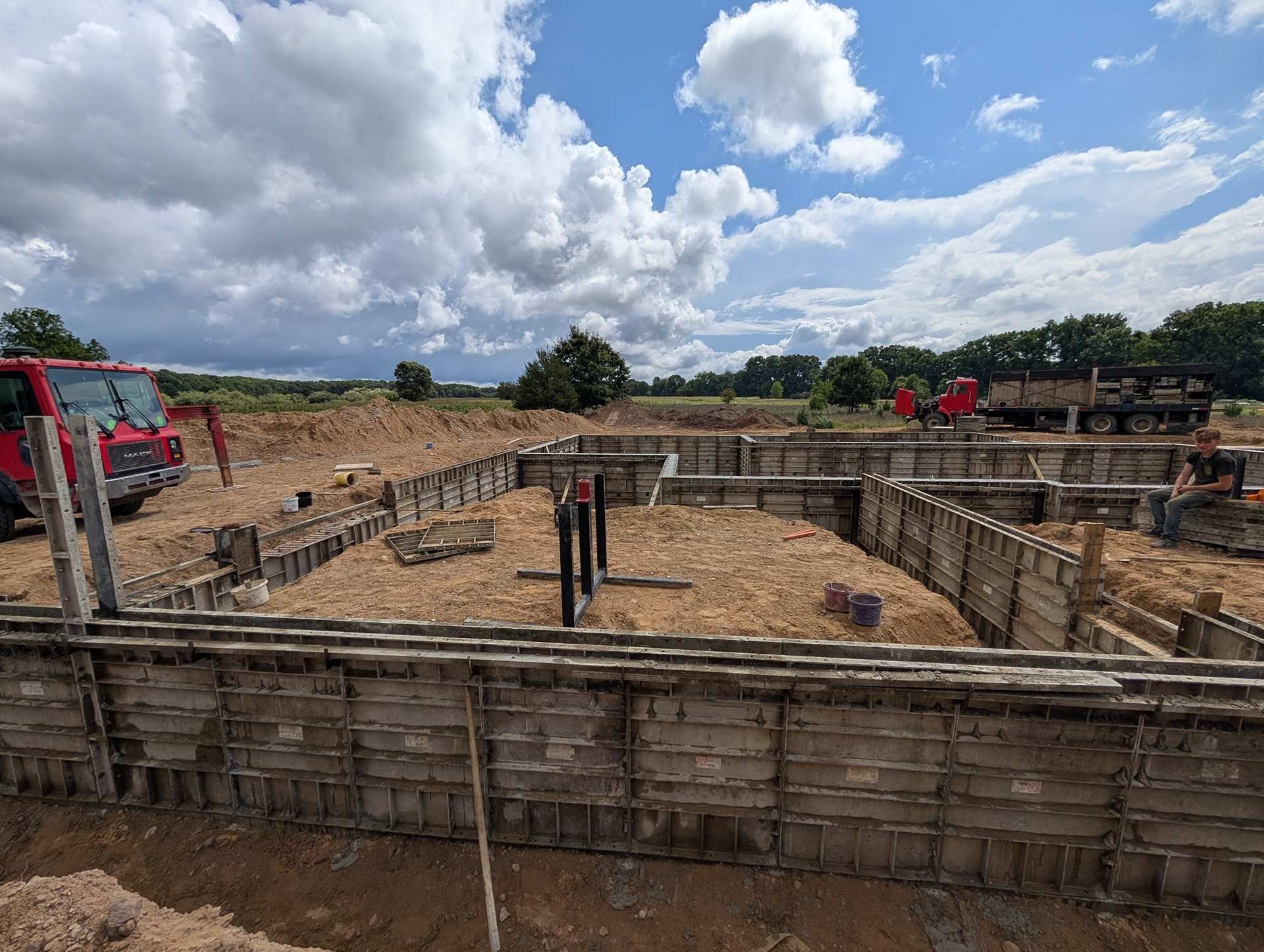 A construction site features concrete foundation forms set in a dirt field under a cloudy blue sky, with vehicles nearby.