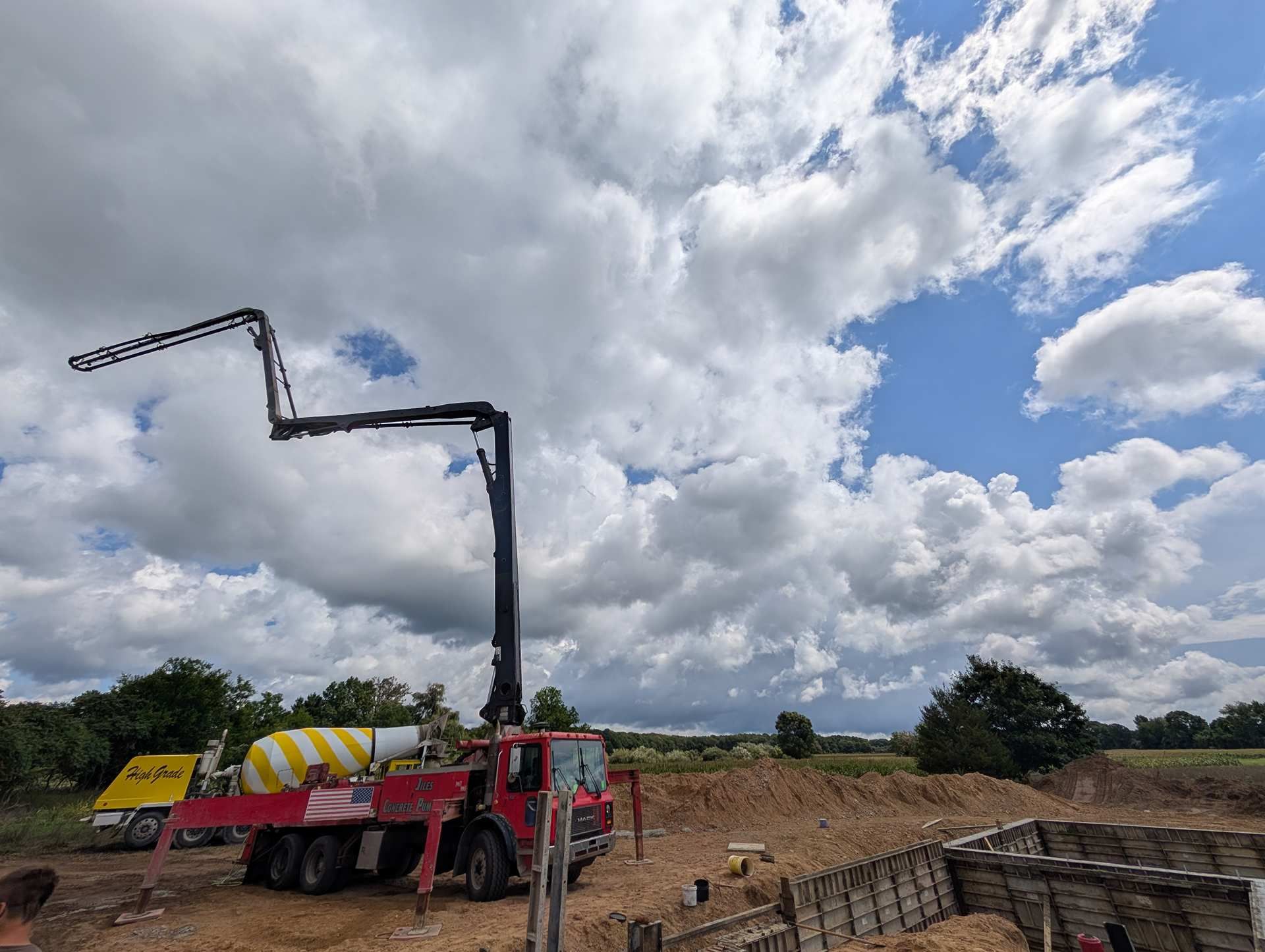 A red concrete pump truck with an extended boom arm, parked at a construction site under a bright, cloudy sky.