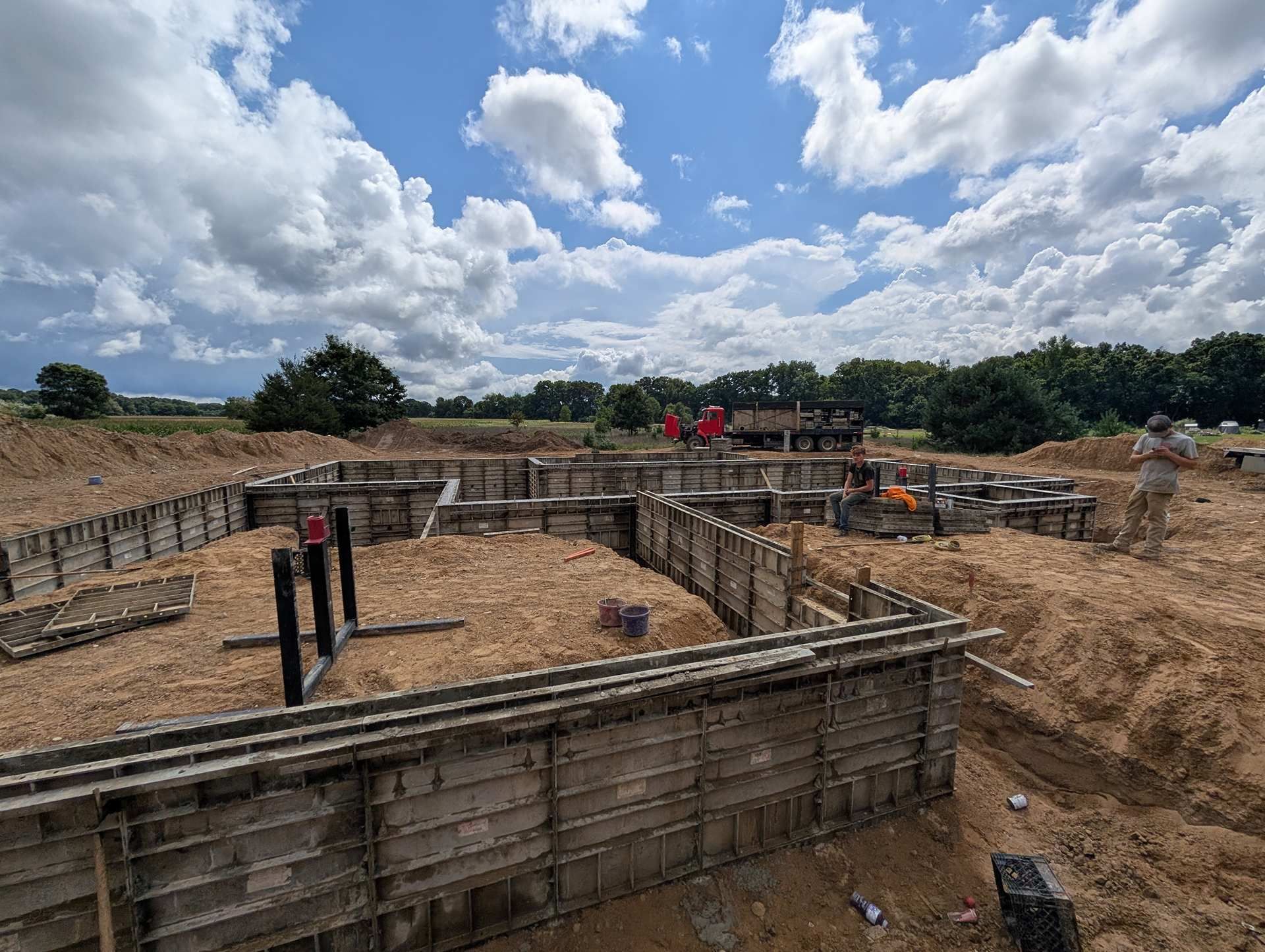 Construction site showing foundation footings with concrete forms set in dirt under a bright, cloudy sky.