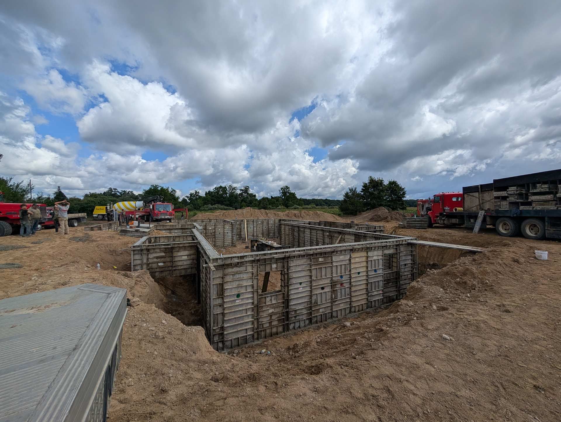 Construction site with concrete formwork for a foundation in a dirt pit under a partly cloudy sky.
