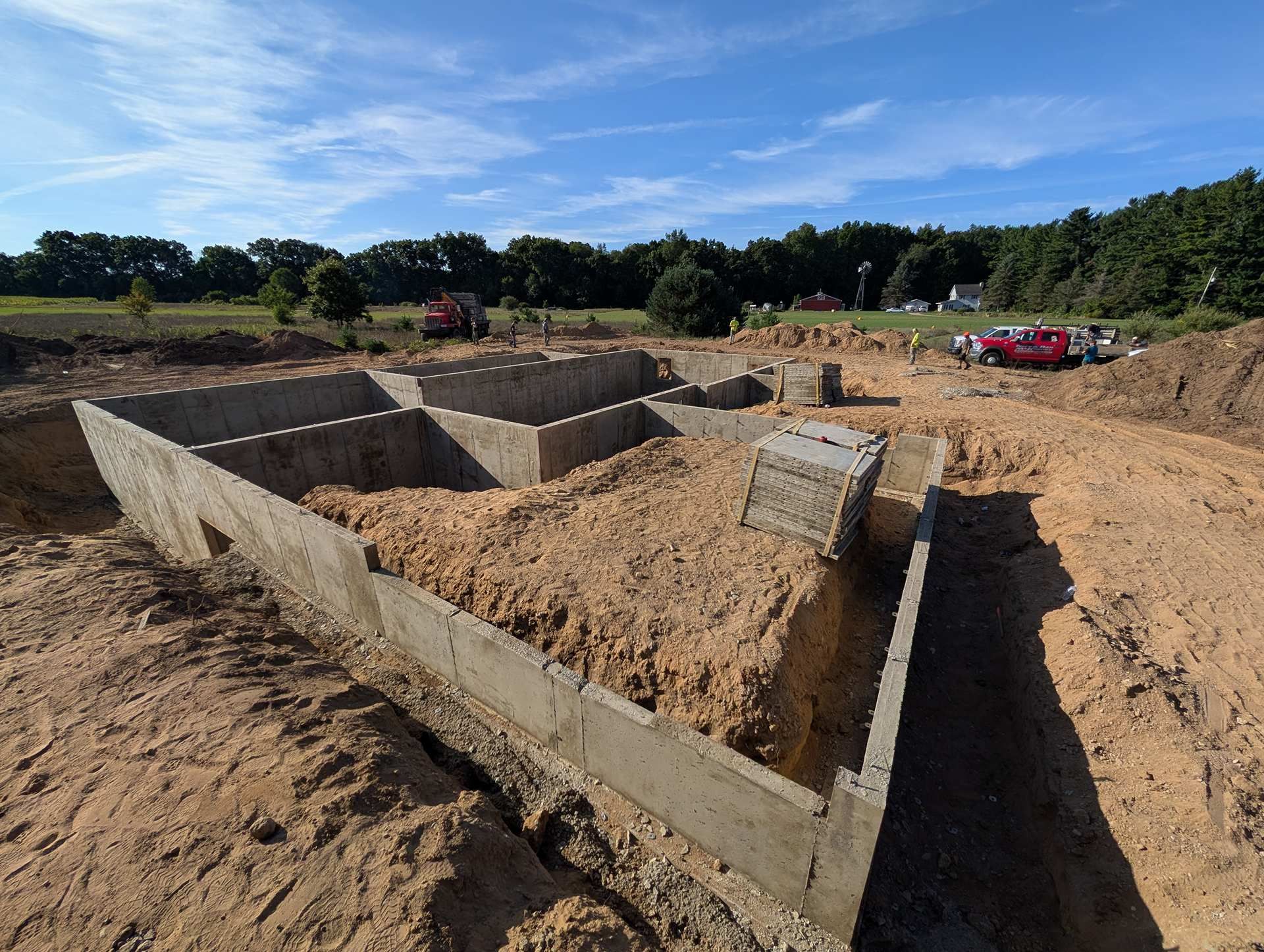 A concrete house foundation under construction on a sunny, rural plot of land with a treeline in the background.