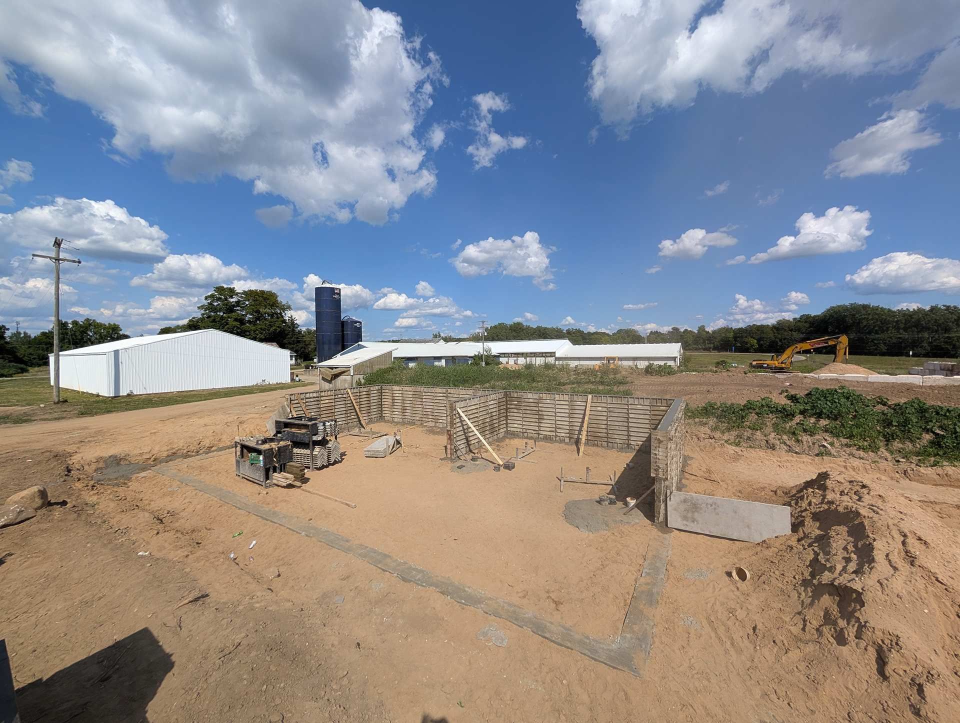 A construction site showing a concrete foundation under a blue, cloudy sky, with industrial farm buildings in the distance.