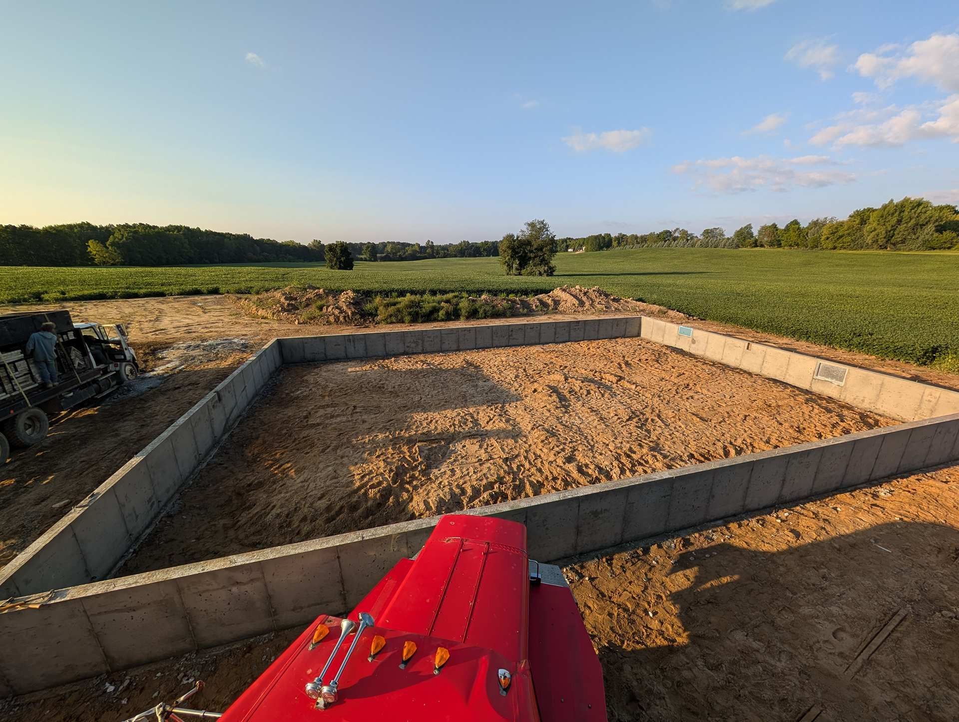 A concrete foundation for a building sits in a rural field with a bright red tractor hood visible in the foreground.