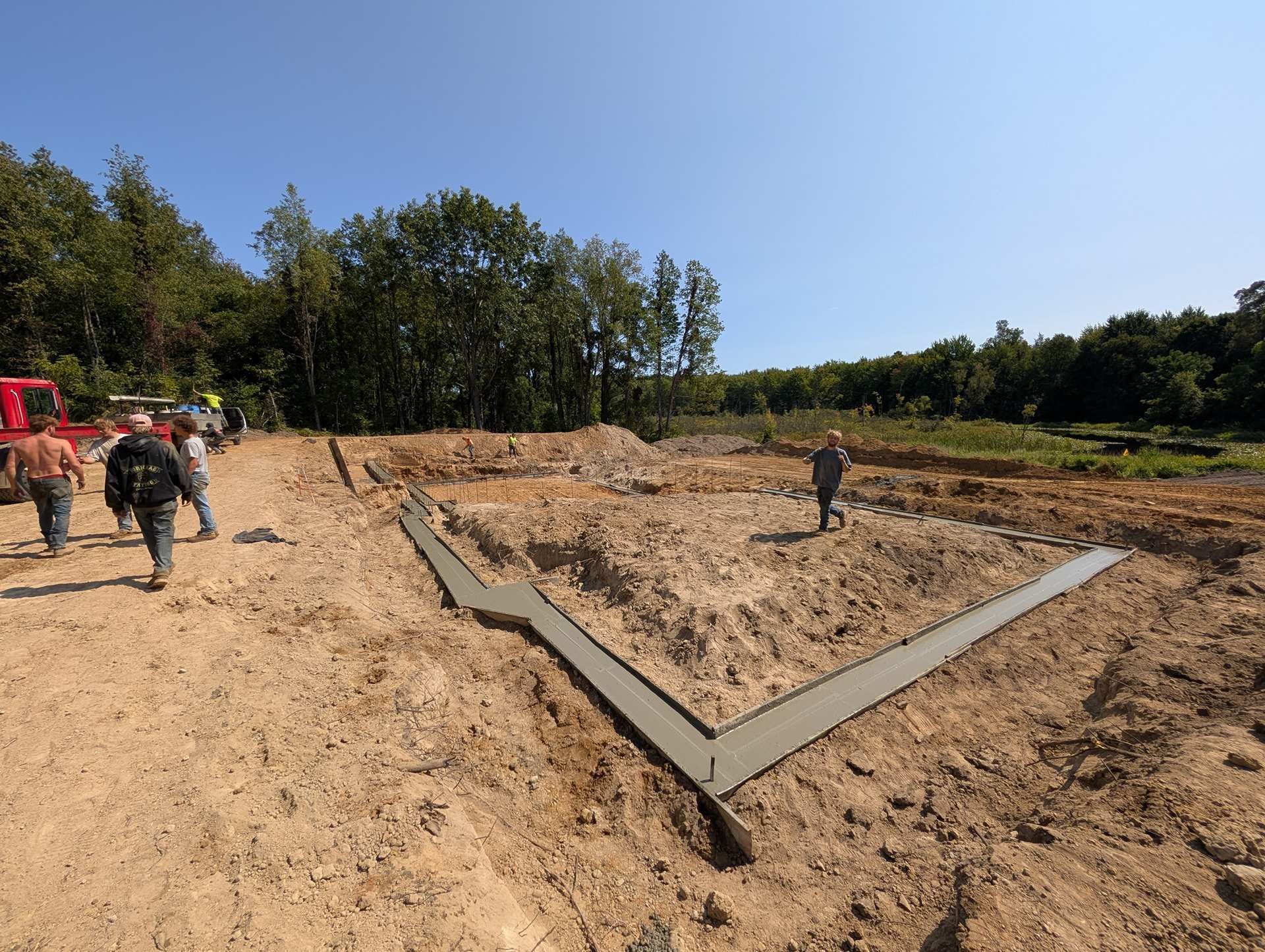 A partially poured concrete foundation for a building in a dirt-filled construction site with workers nearby.