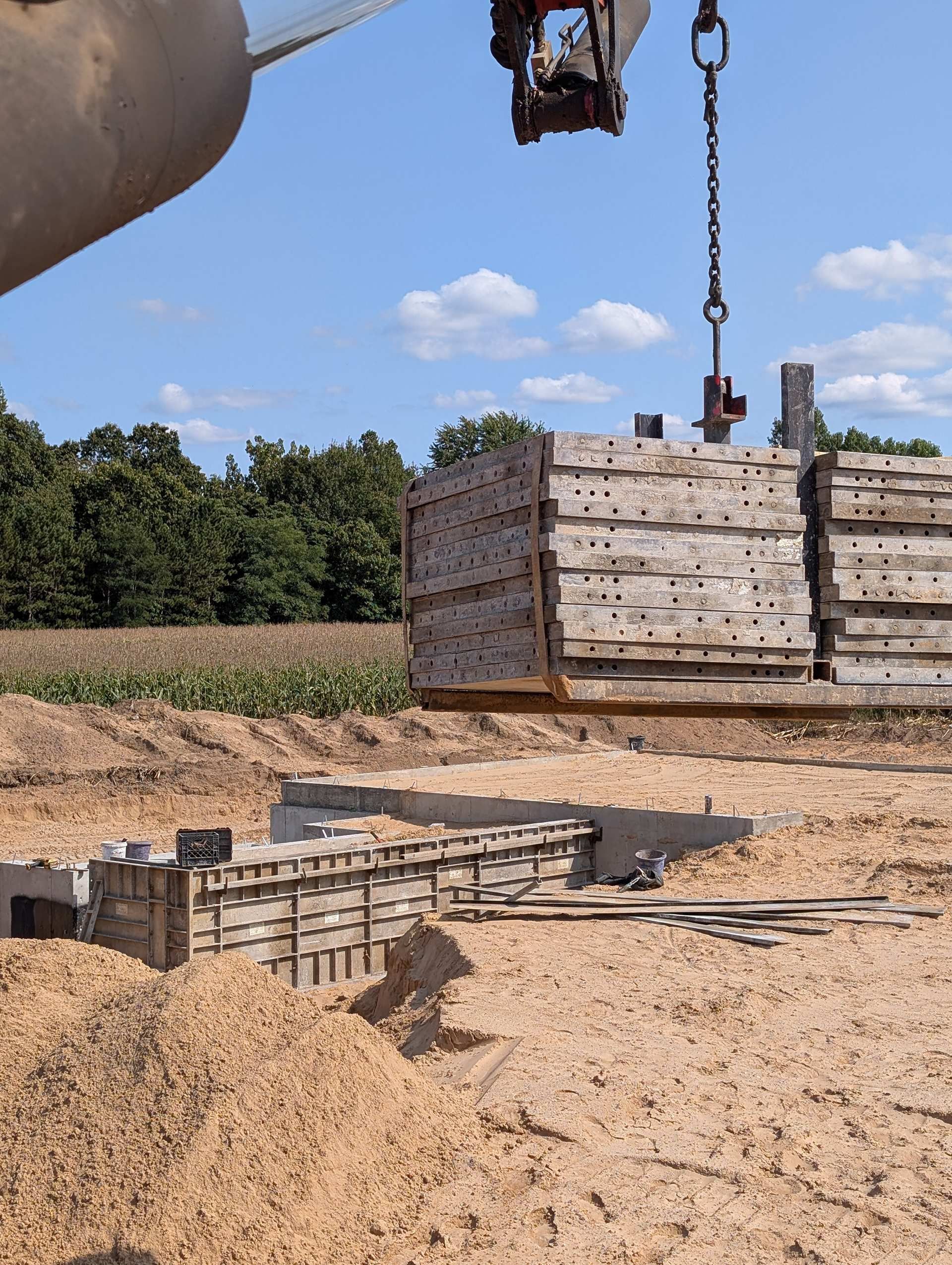 A construction crane lowers a stack of heavy concrete formwork panels onto a dirt building site.