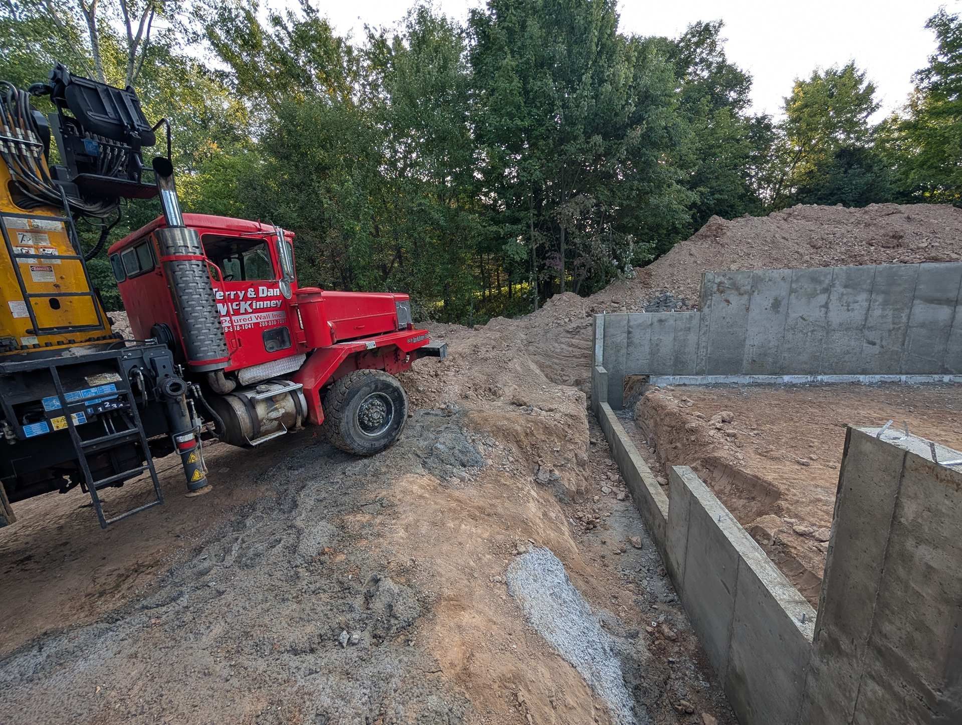 A red utility truck sits on a dirt construction site next to a concrete foundation wall with trees in the background.