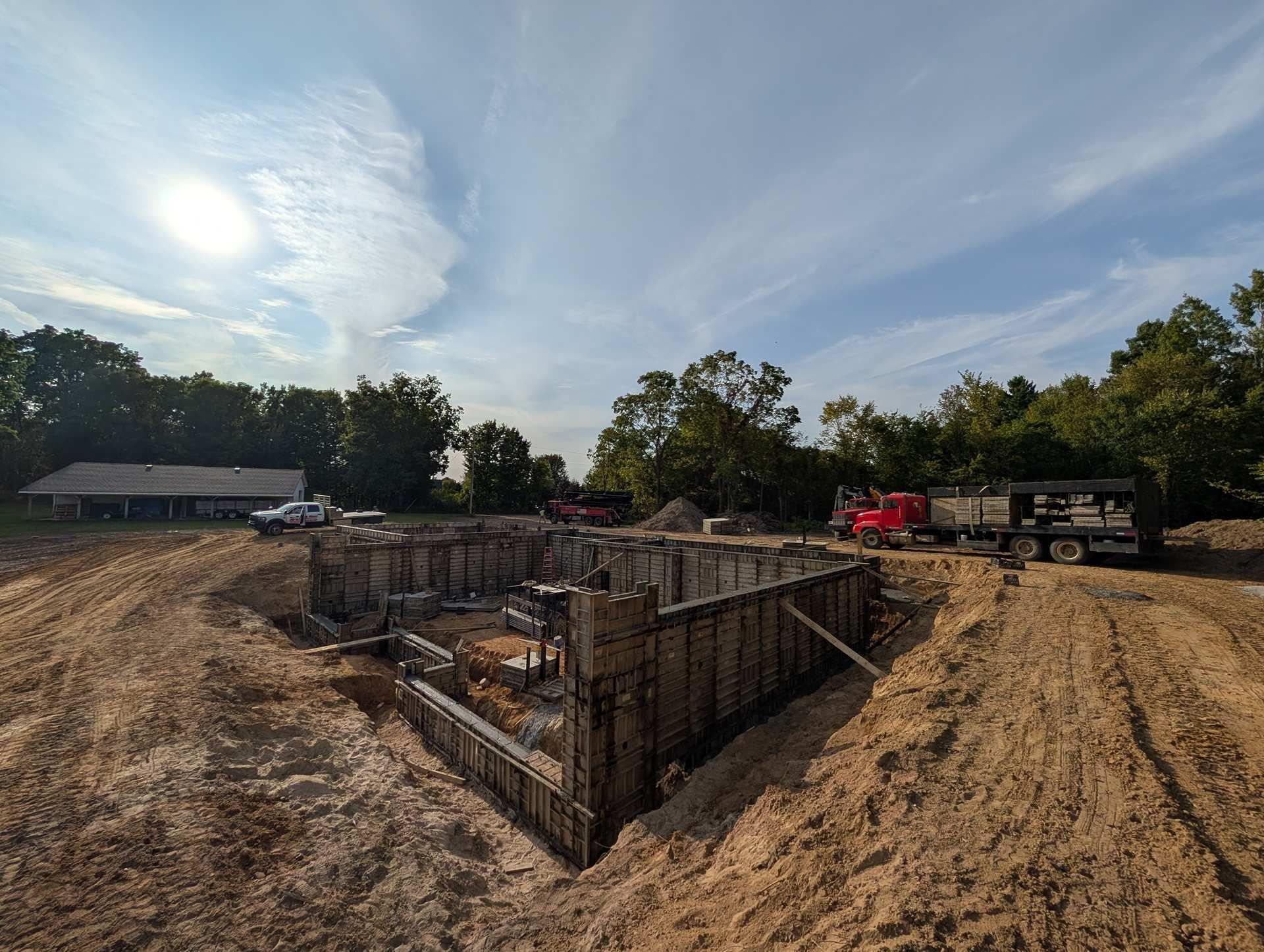 A foundation with concrete forms under construction at a sunny, rural job site with construction trucks nearby.