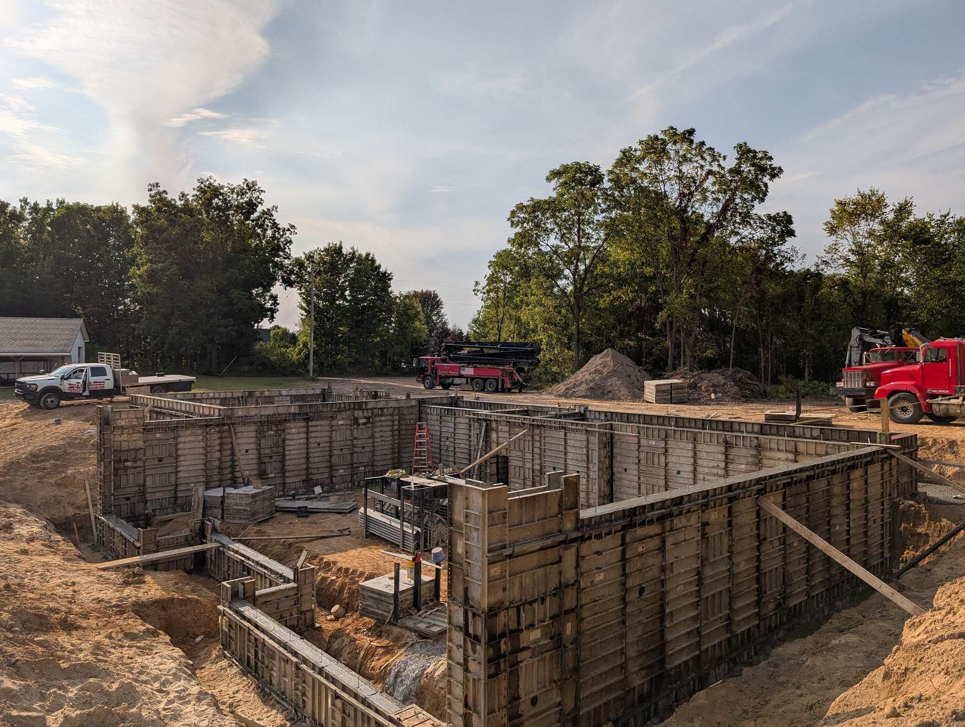 Concrete foundation forms installed in an excavated pit at a construction site with trees and equipment in the background.