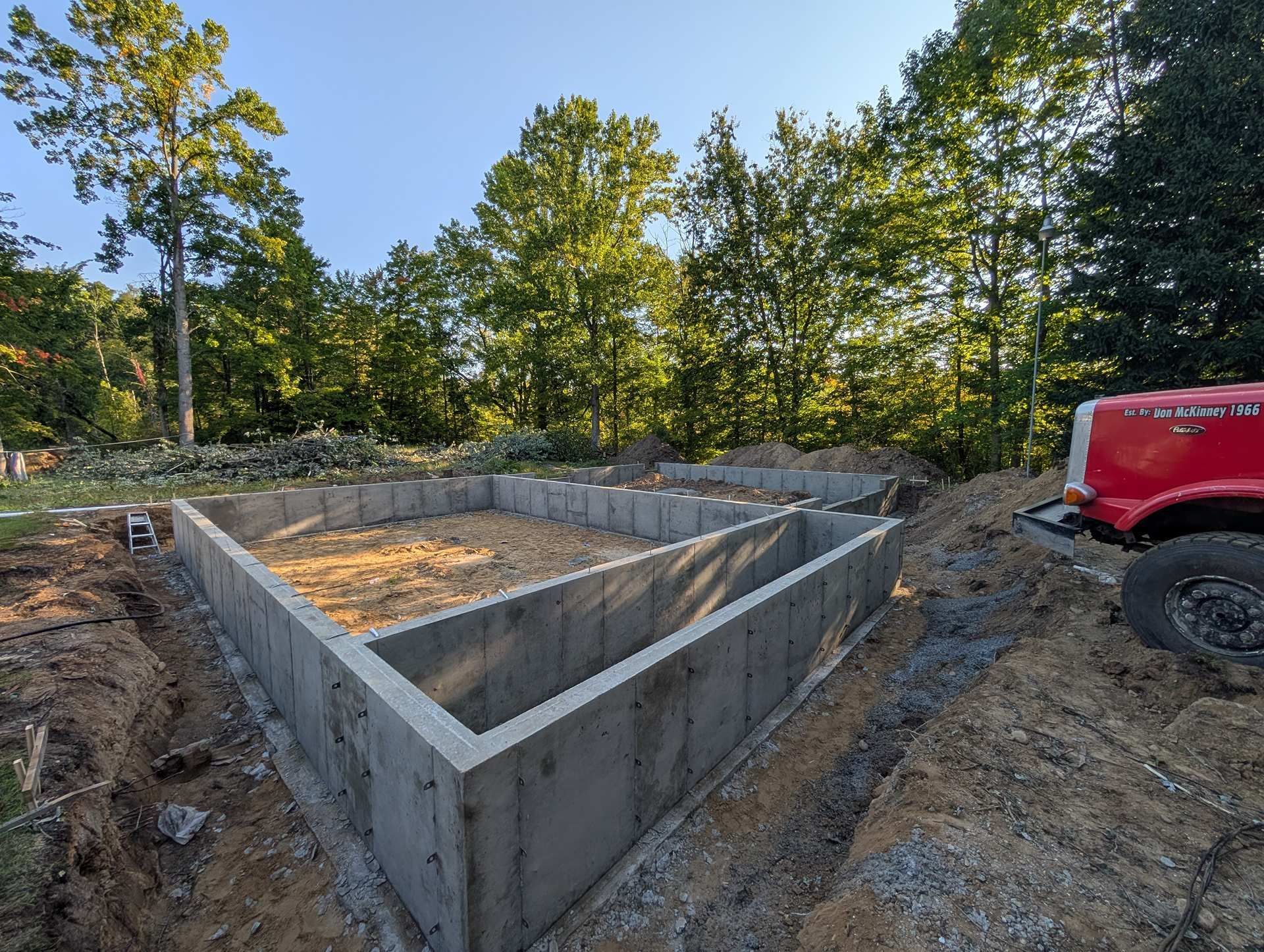 Concrete foundation walls for a new house under construction, situated in a wooded area next to a red truck.
