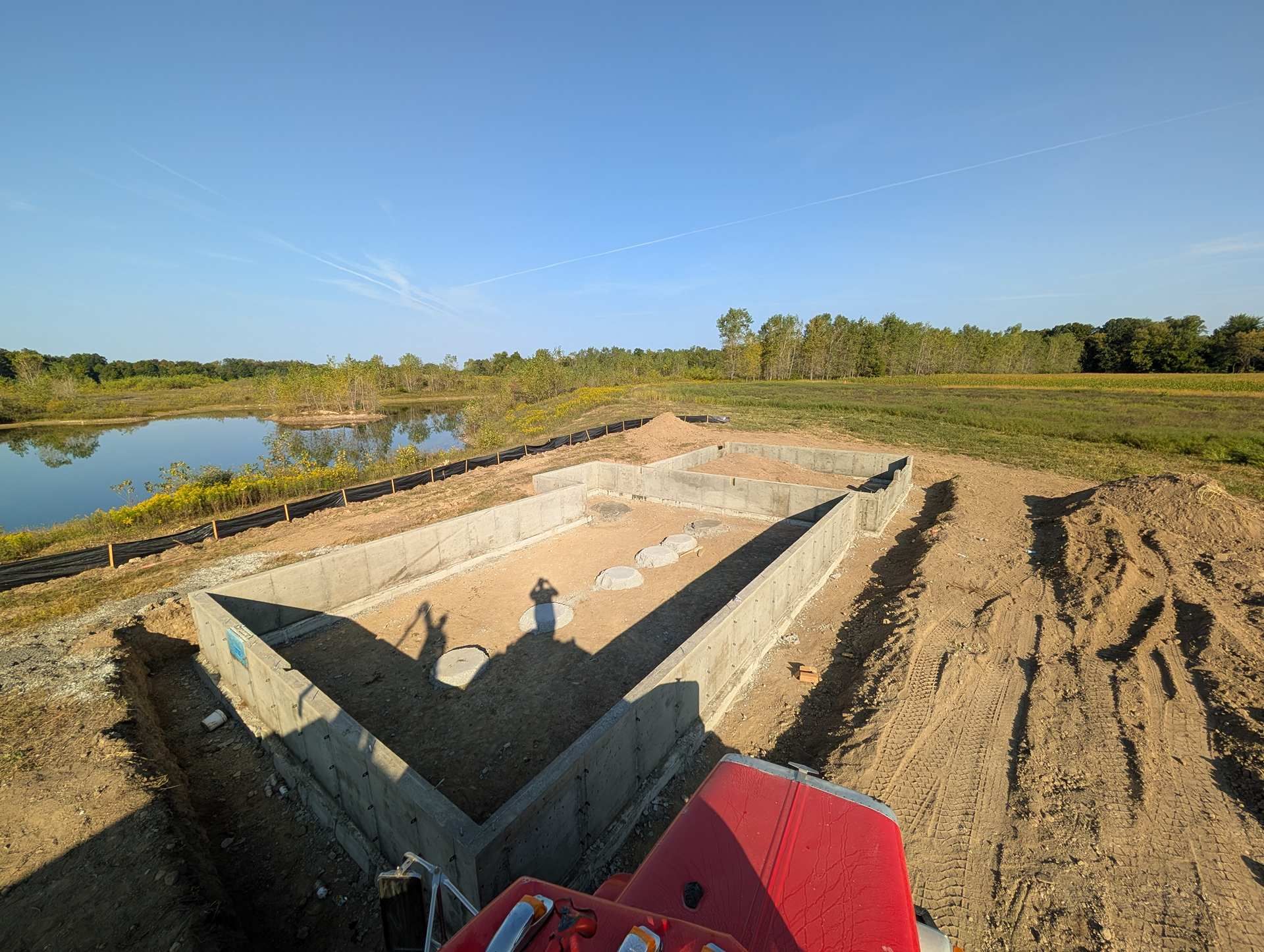 A concrete foundation for a building under construction, overlooking a pond in an open, grassy field under a blue sky.